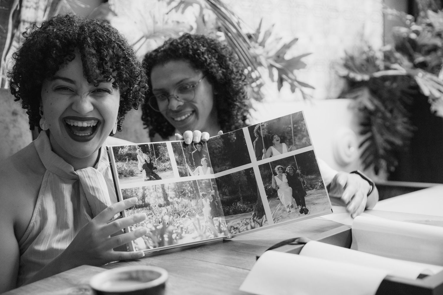 Two women with curly hair laugh while holding open a photo album at a cafe table, captured in black and white.