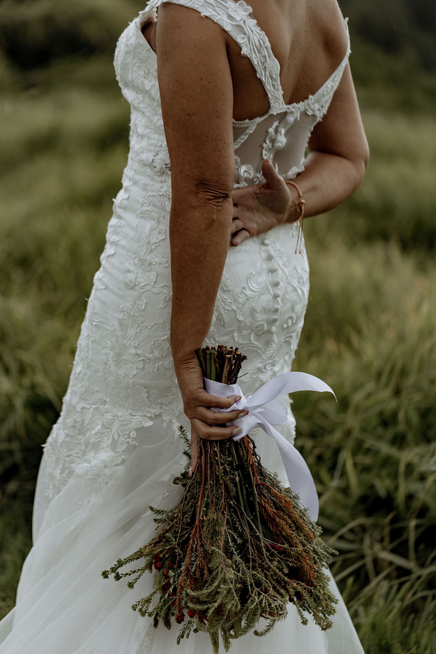 A bride in a white lace wedding dress holds a rustic dried flower bouquet against a grassy outdoor backdrop.