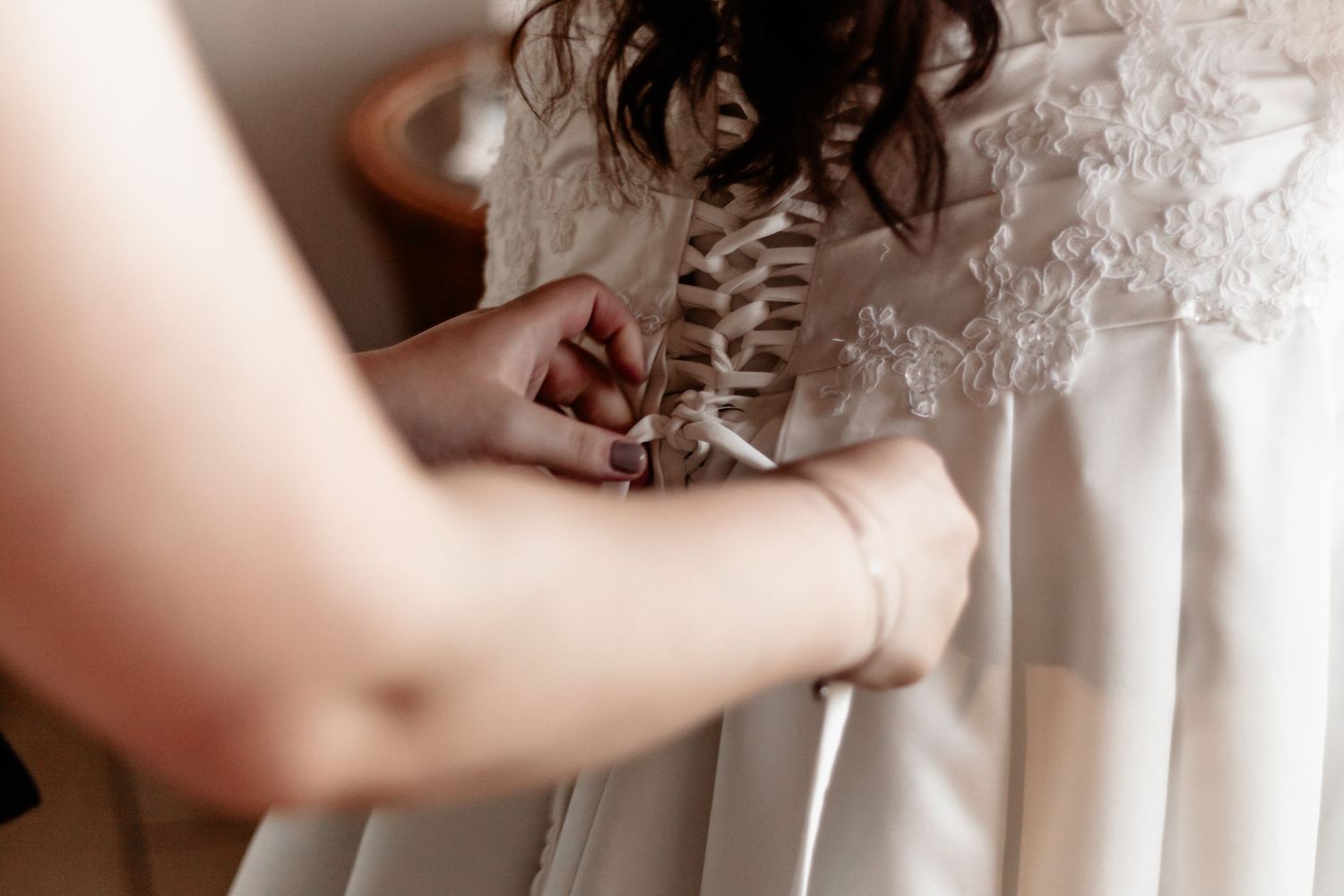 Close-up view of hands fastening corset laces on the back of a white wedding dress.