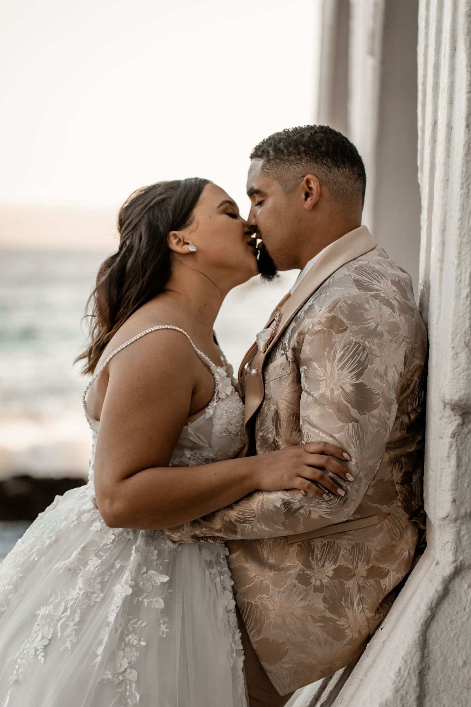 Romantic wedding couple sharing an intimate moment at sunset by a white pillar in elegant floral attire.