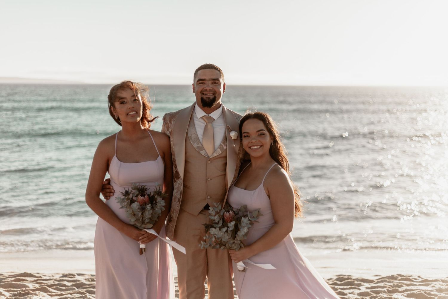 Wedding party posing together on a sunny beach with bouquets and formal attire as waves roll in behind them.