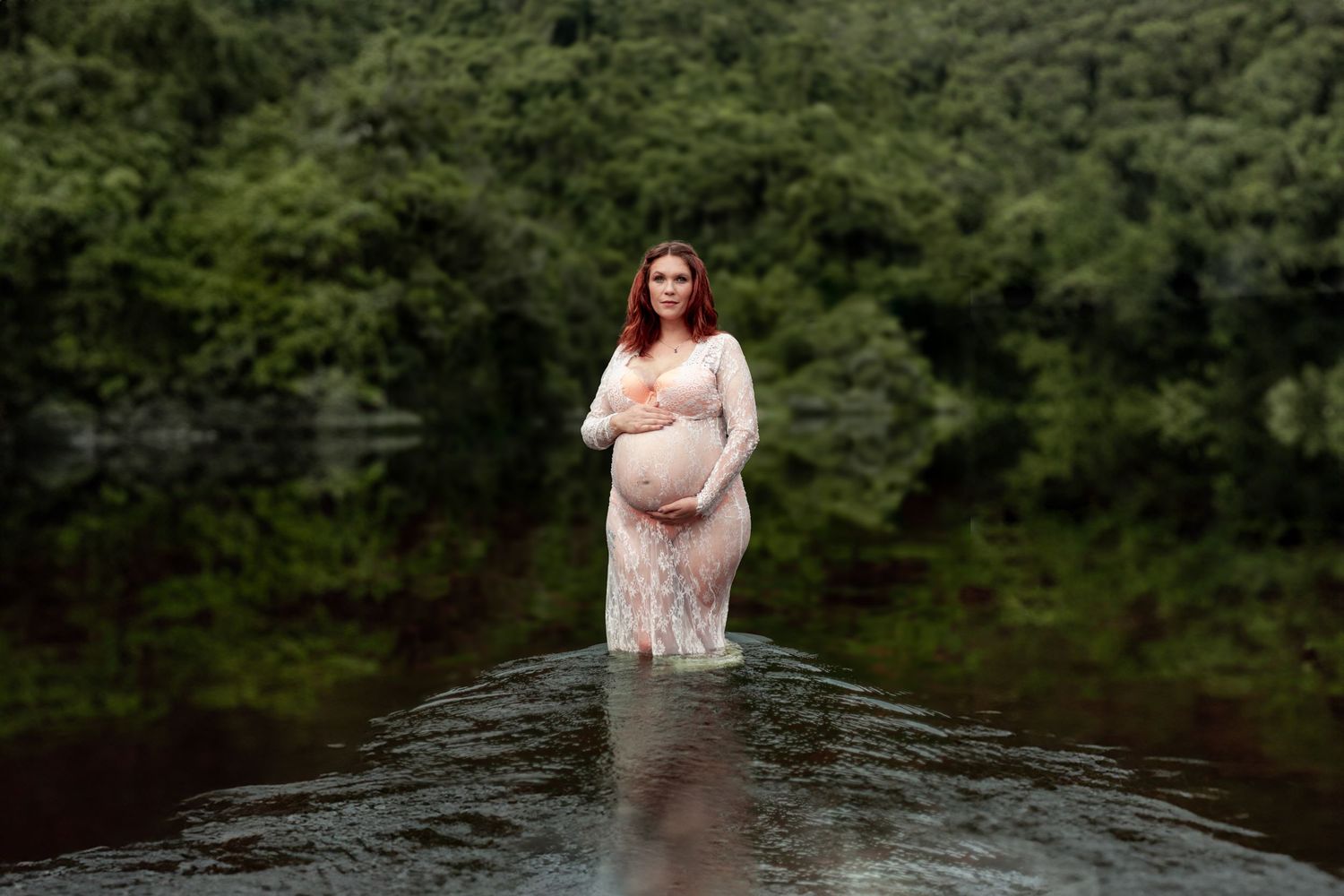 A pregnant figure in a pink gown poses in a wooded area near dark water for maternity photos.