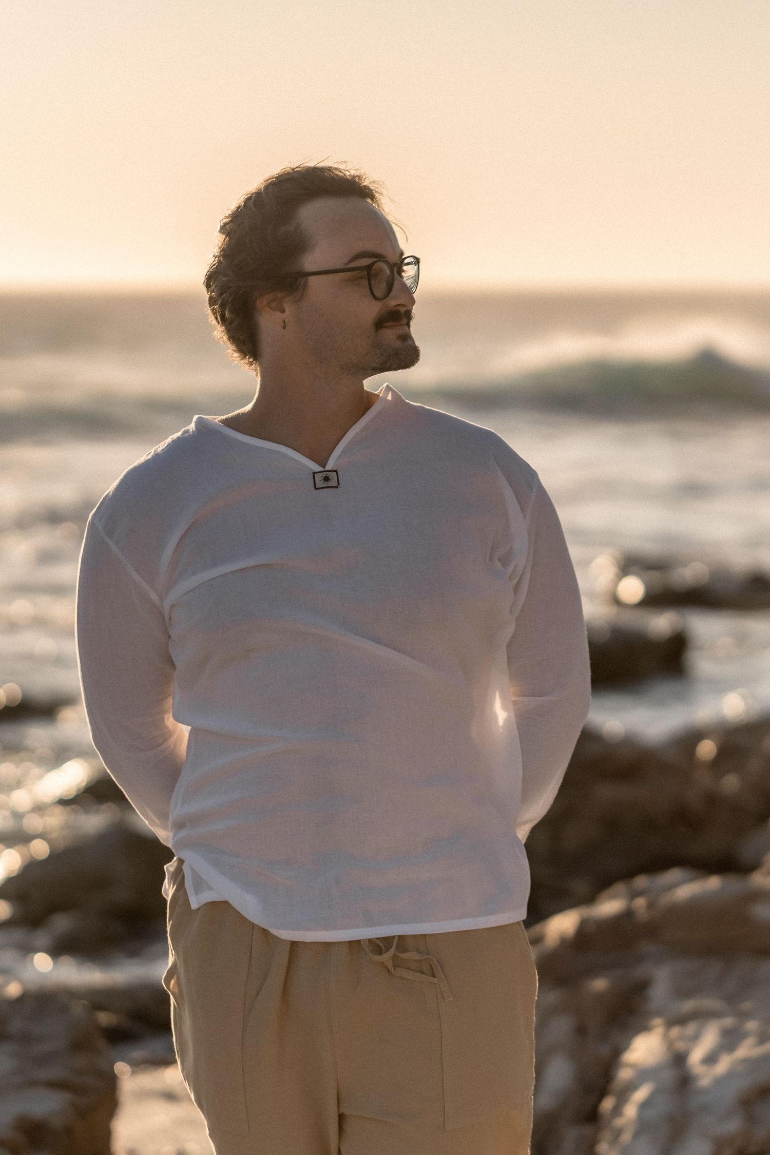 Person wearing a white sweater and khaki pants stands contemplatively by the ocean at sunset.