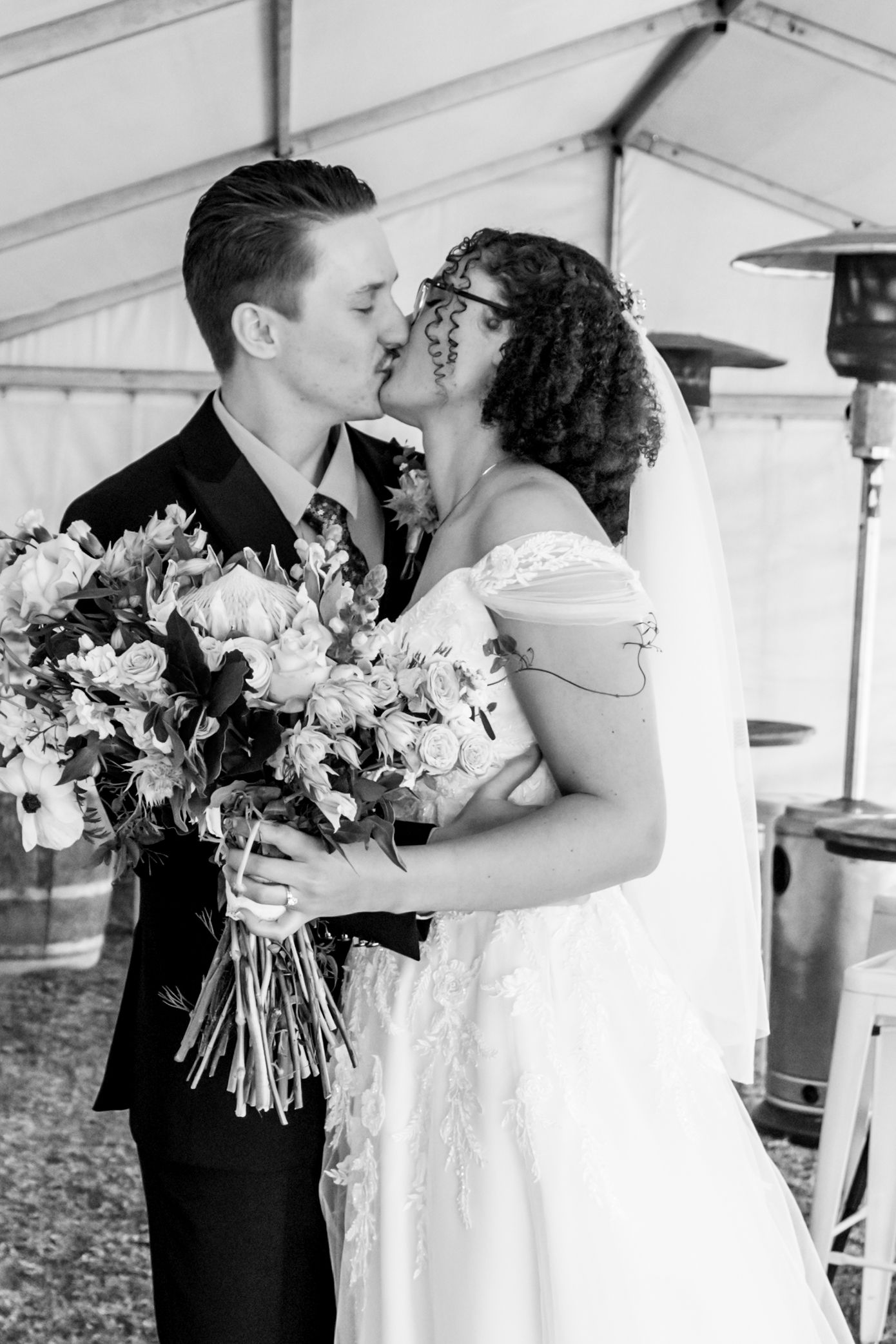 A romantic wedding couple shares tender moments and kisses under a white tent with floral bouquets.