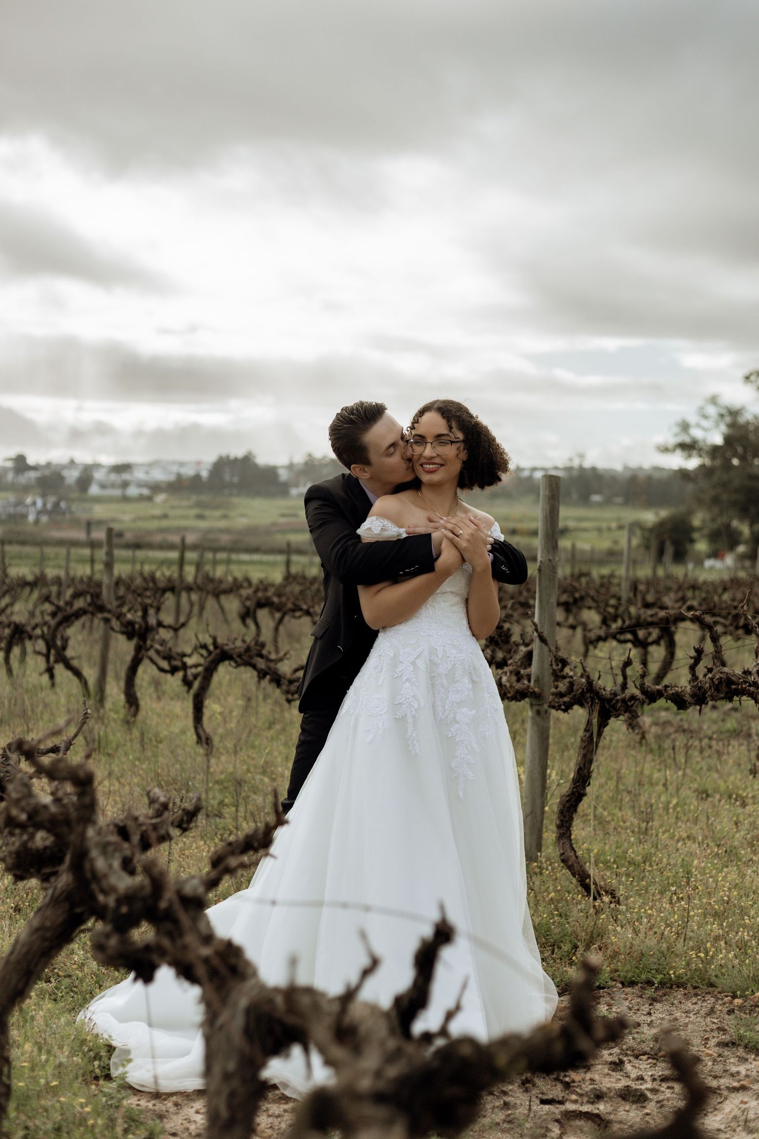 A couple in wedding attire shares an intimate dance among old grapevines in a rustic vineyard setting.