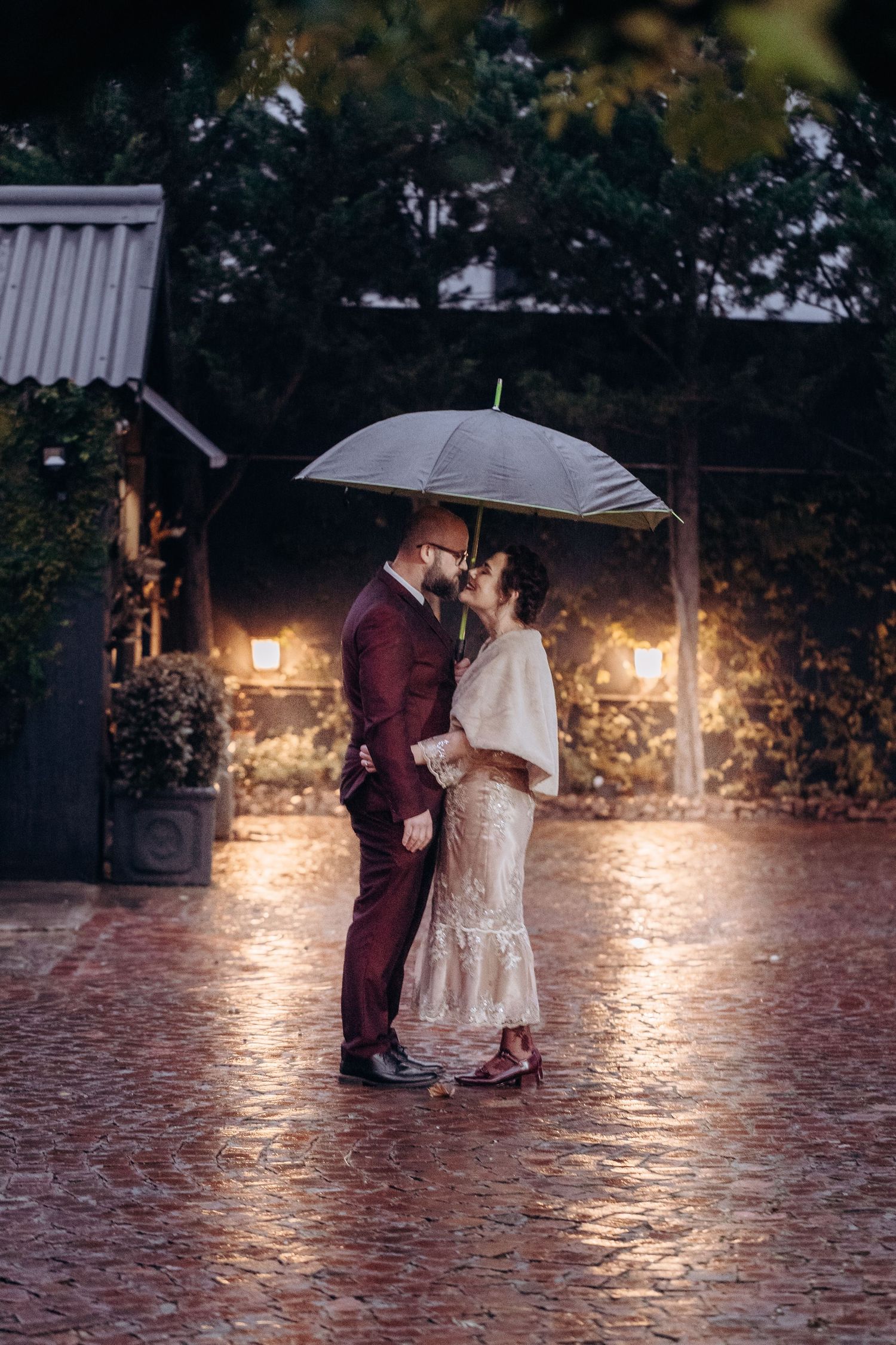 Couple sharing a romantic kiss under a clear umbrella on a rainy evening, groom in burgundy suit, bride in white lace dress.