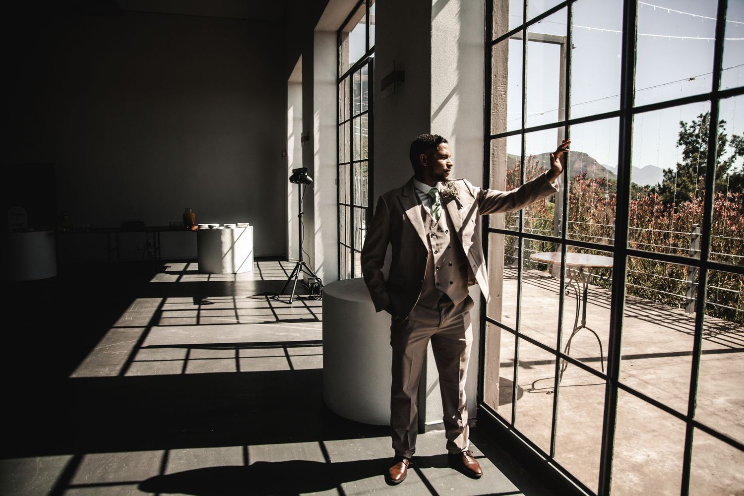 A well-dressed Black man in a tailored suit stands by large industrial windows in a sunlit modern space.