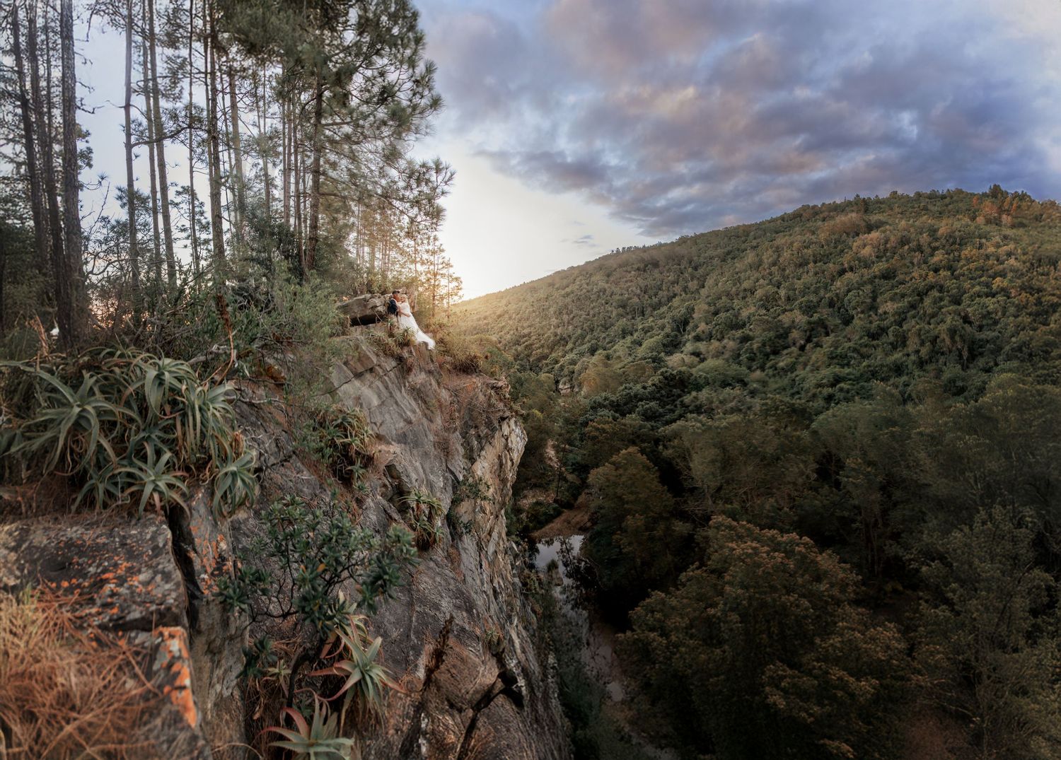 Scenic rocky cliff overlooking a lush green forested valley with a waterfall and dramatic sunset sky.