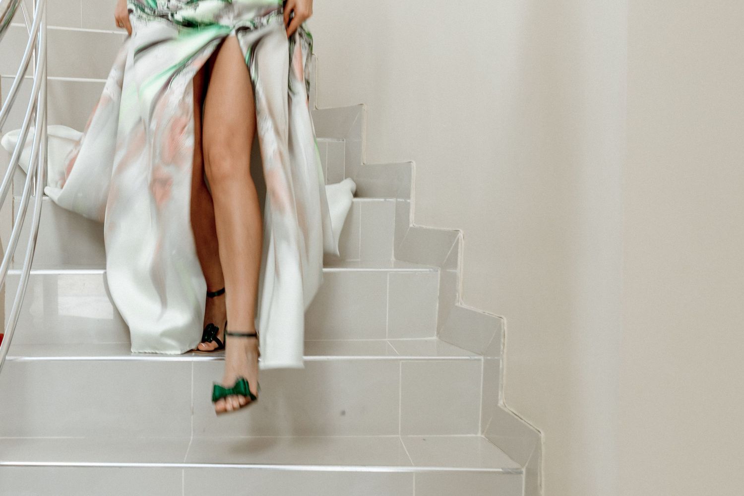 Legs in elegant green heels descending white stairs while wearing a flowing light-colored dress.