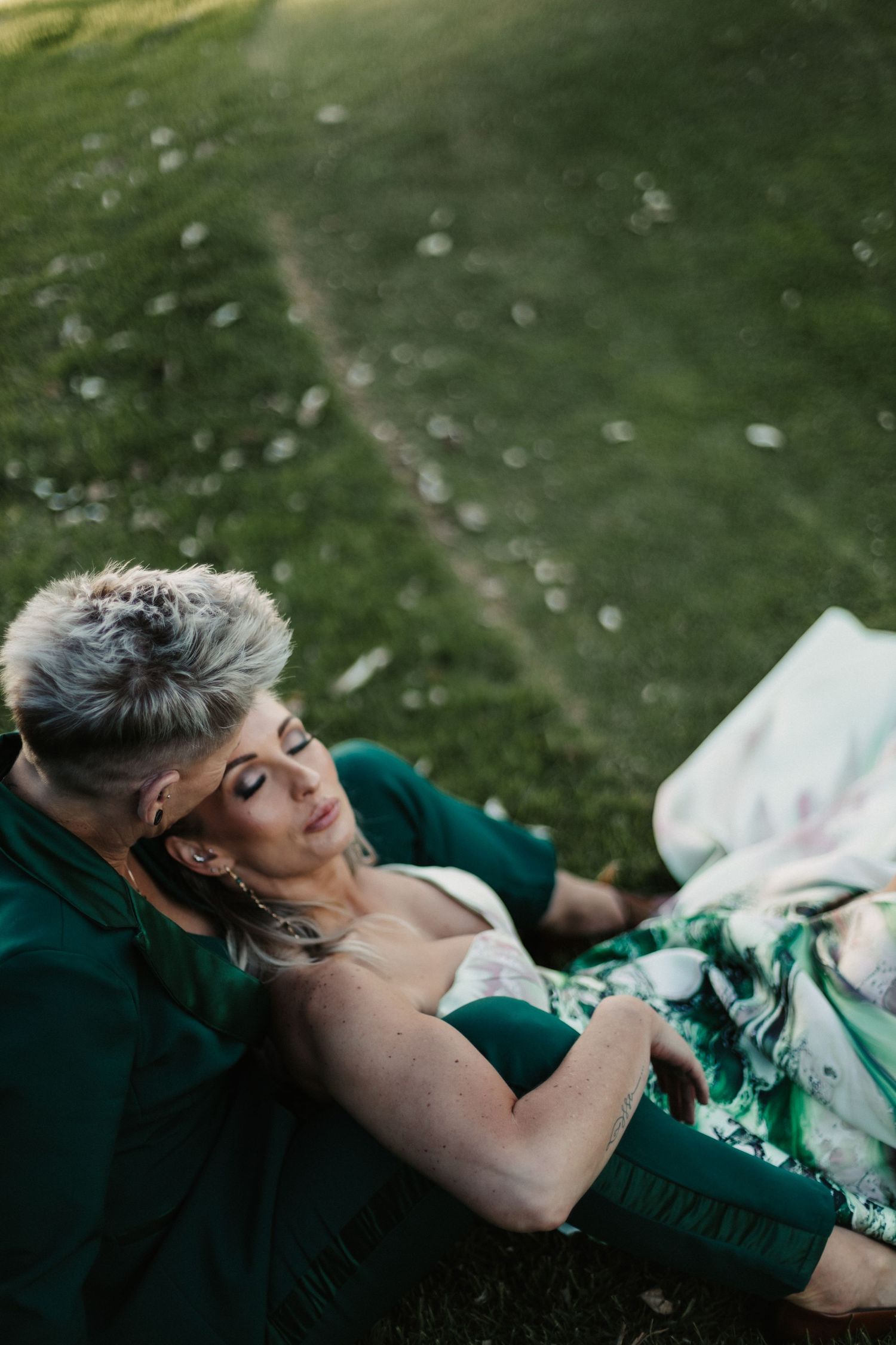 Person in an emerald green dress lies gracefully on grassy lawn during an outdoor photoshoot.
