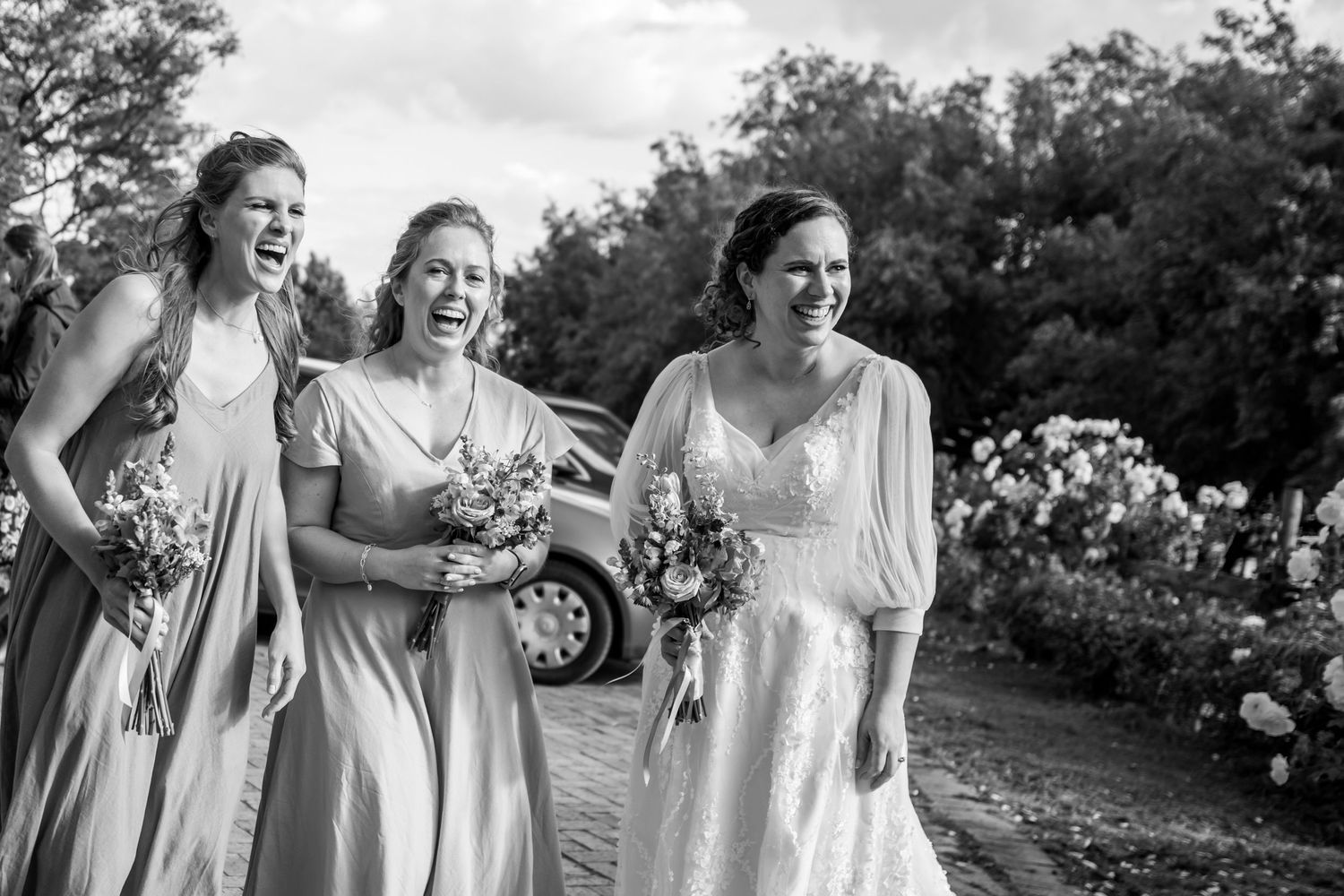 A group of bridesmaids in light-colored dresses laugh and walk together through a garden setting in black and white.