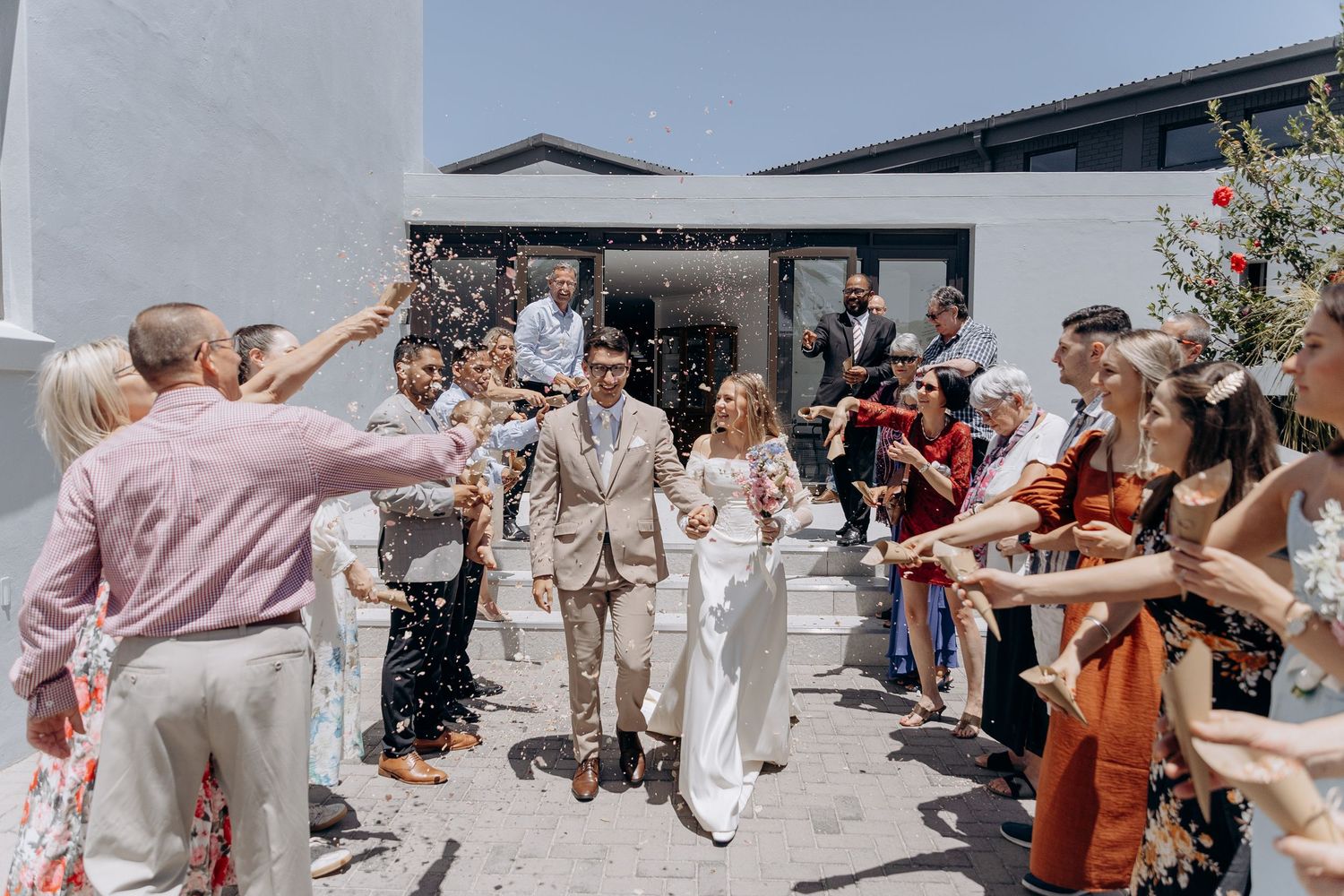 Wedding guests throw flower petals and confetti as the newlyweds walk through a joyful celebration line outdoors.