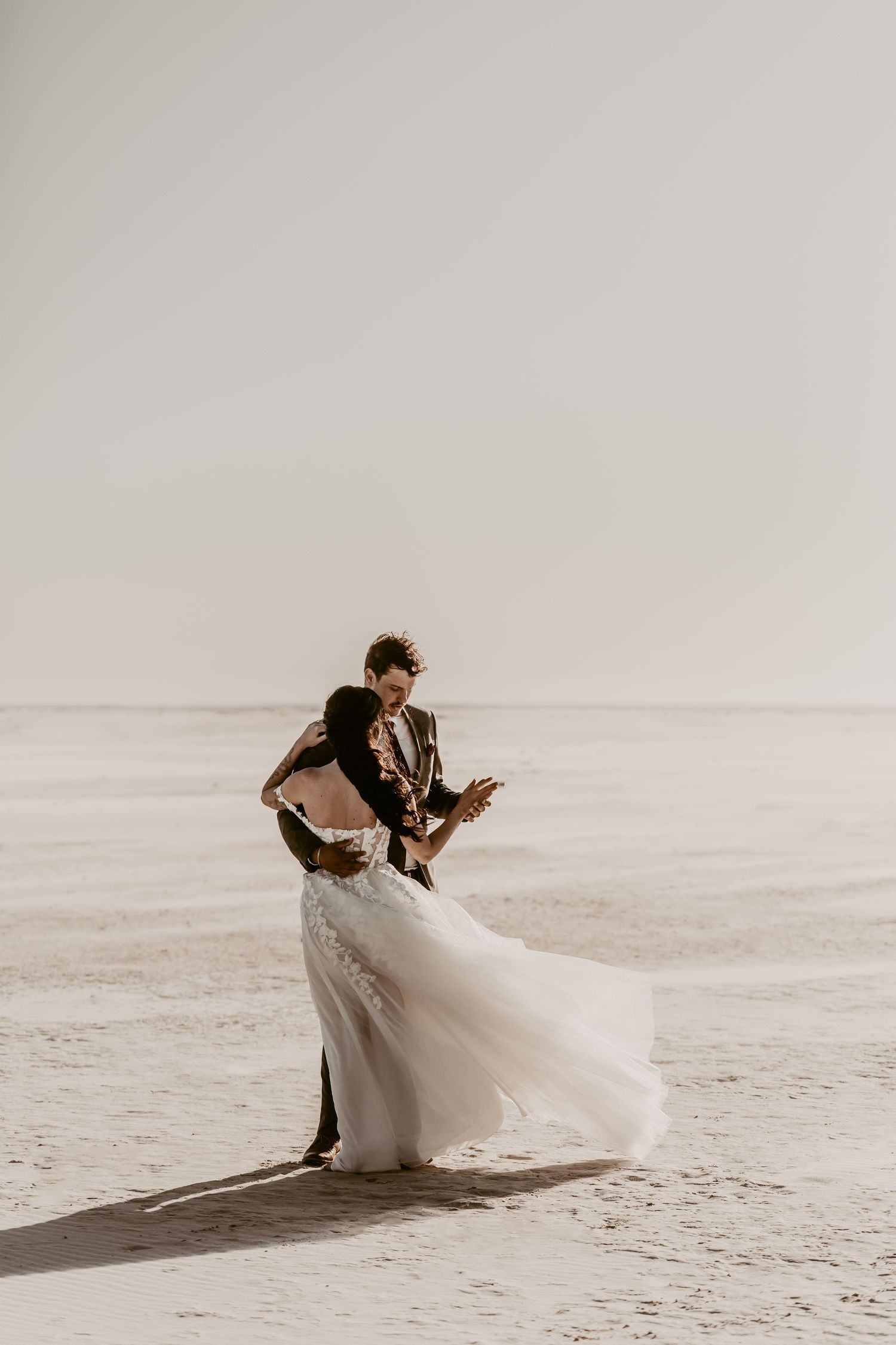 A couple in wedding attire embraces on a misty beach as the bride's white dress flows in the wind.