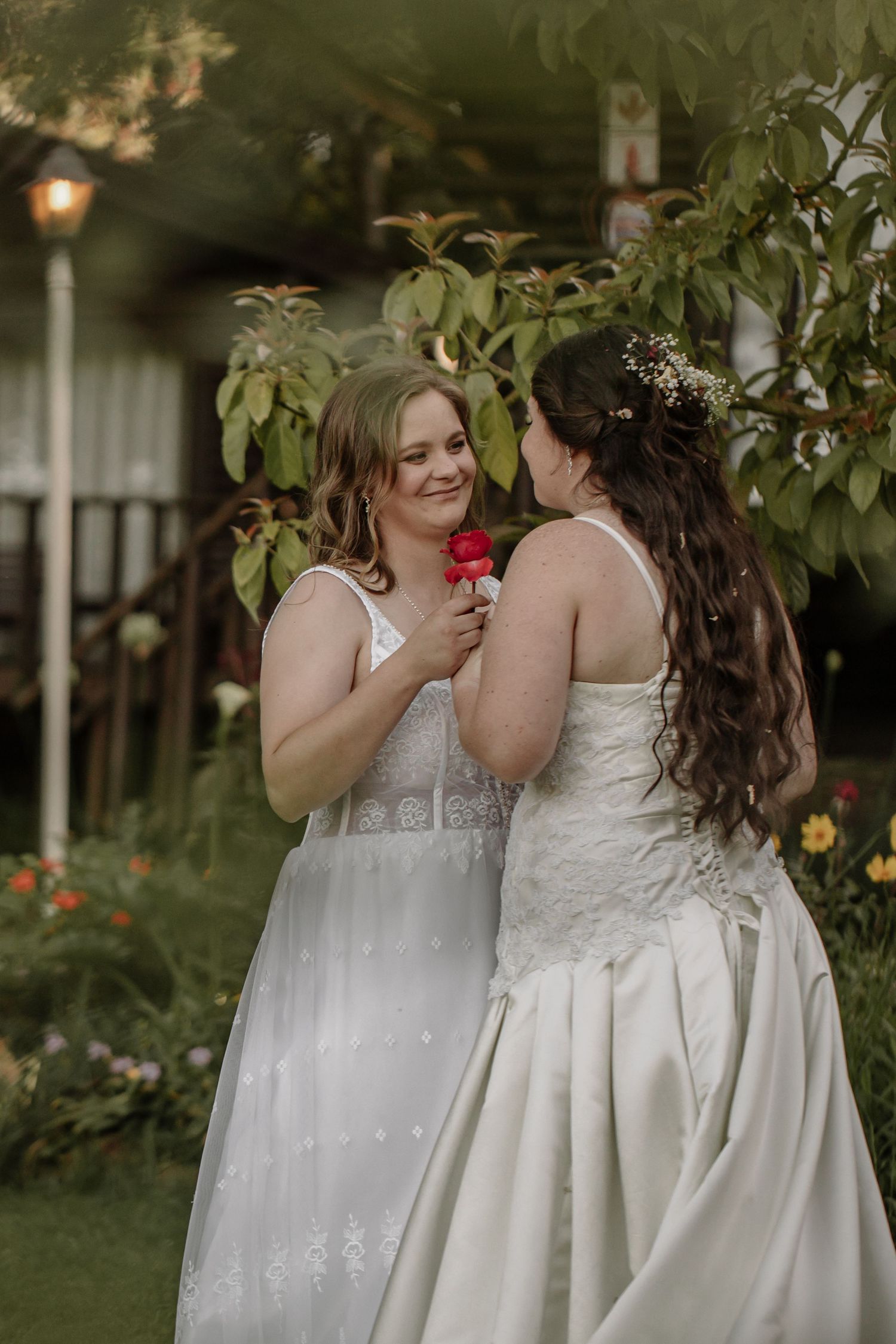 Two brides share an intimate moment in a garden wearing white wedding dresses with floral accents and elegant details.