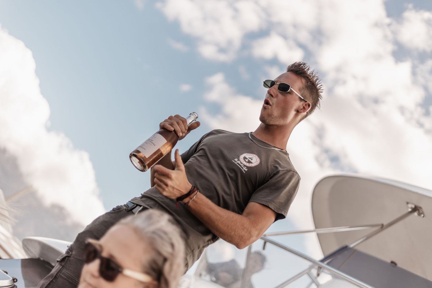 Person in sunglasses holding a beverage while standing on a boat against a cloudy sky background.