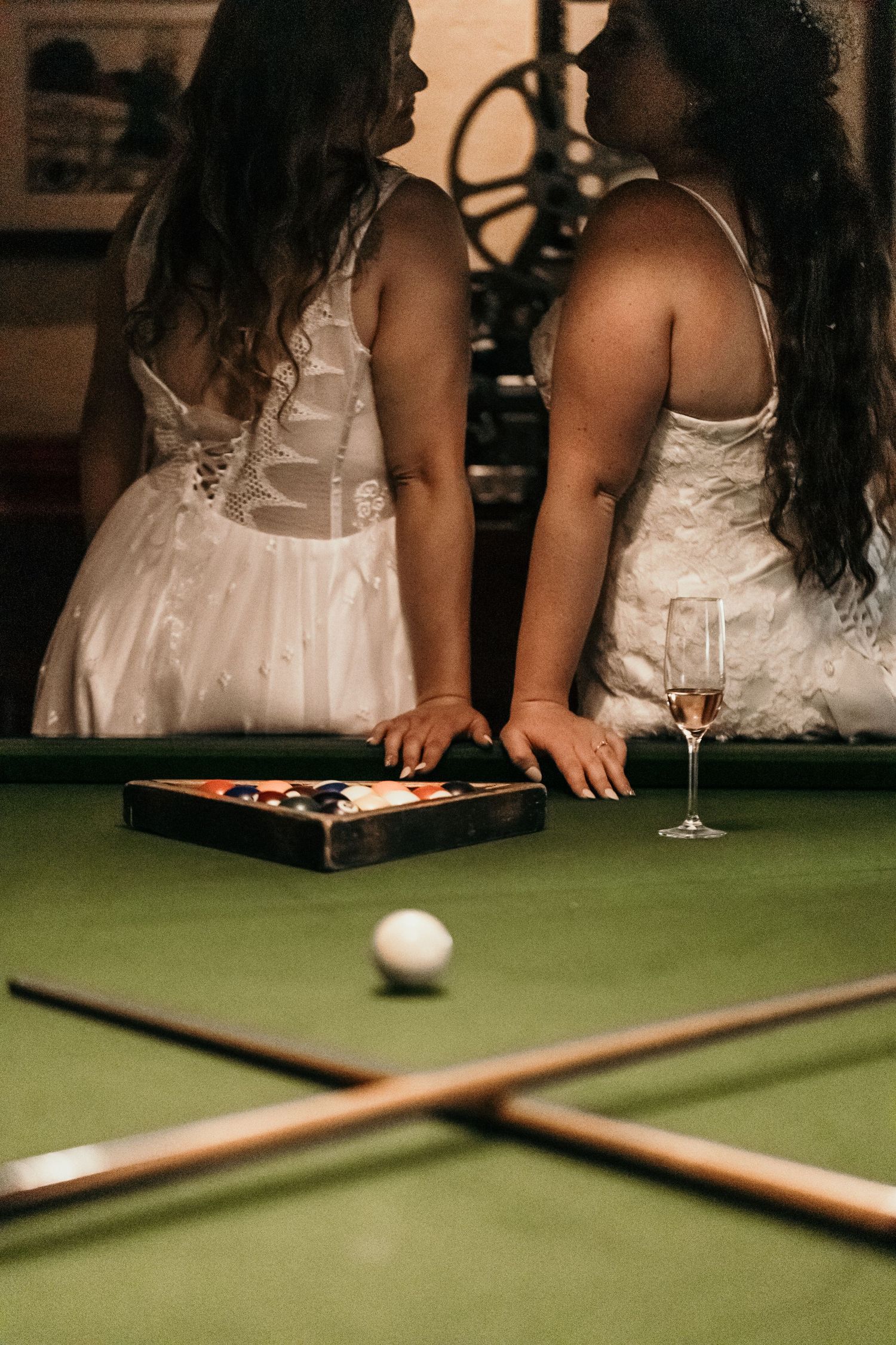 Two people in white dresses play pool while enjoying a glass of wine at an elegantly lit venue.
