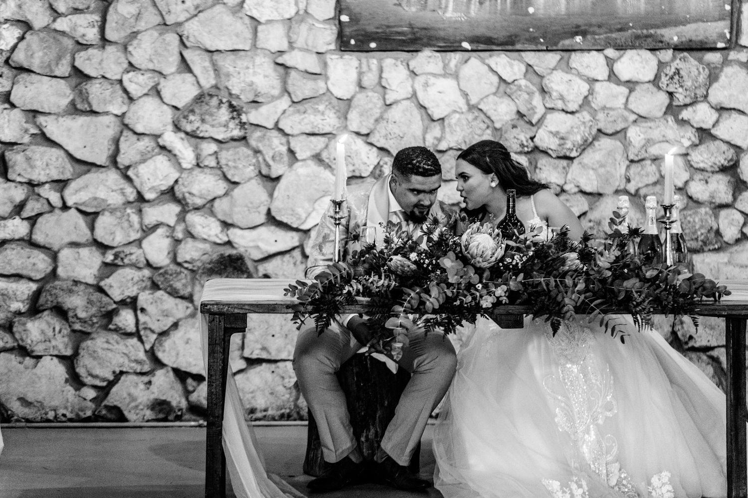 Wedding party celebrates together in front of rustic stone wall backdrop at outdoor reception venue.