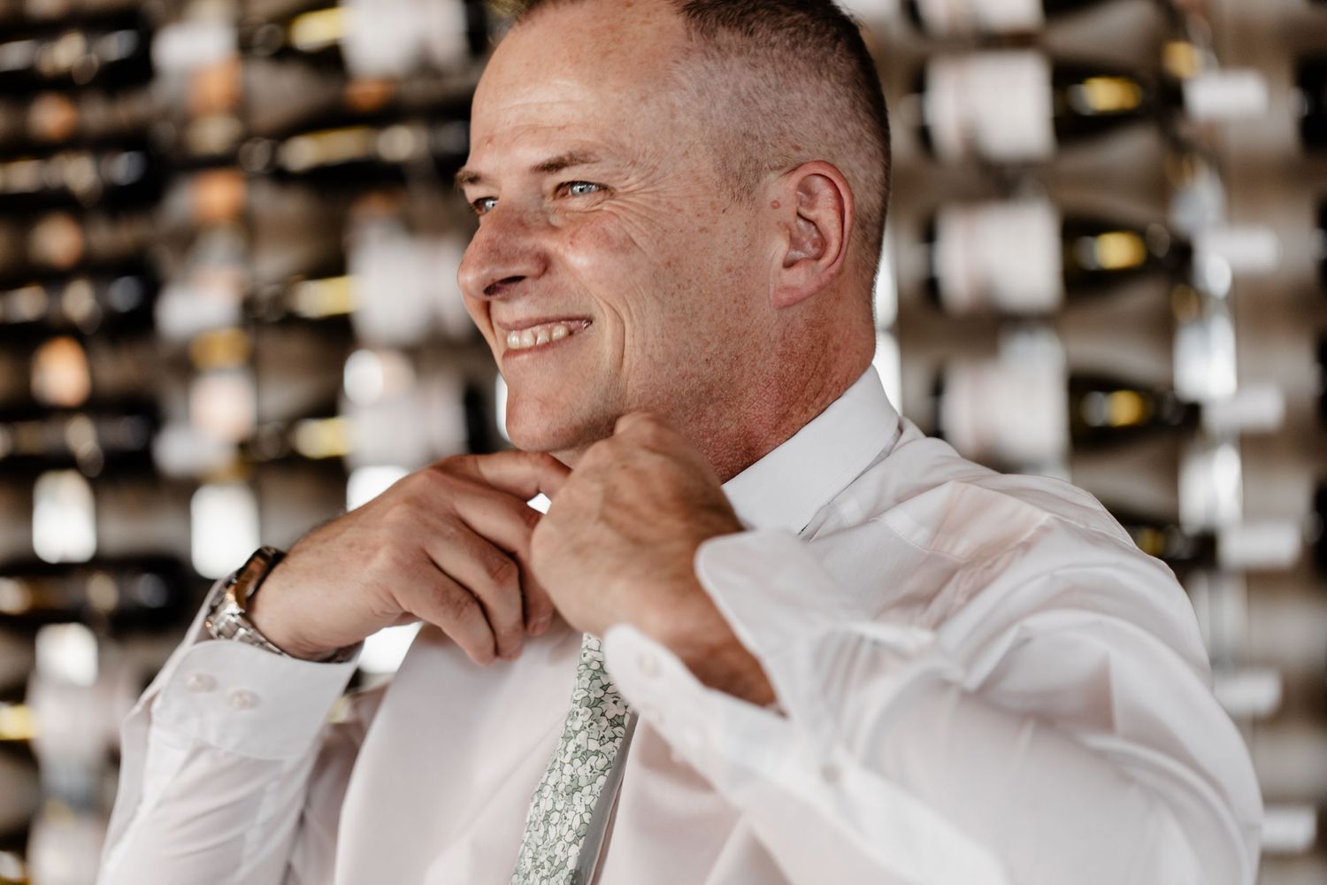 A person in white dress shirt and greenish tie smiles while standing in front of a wall of wine bottles.