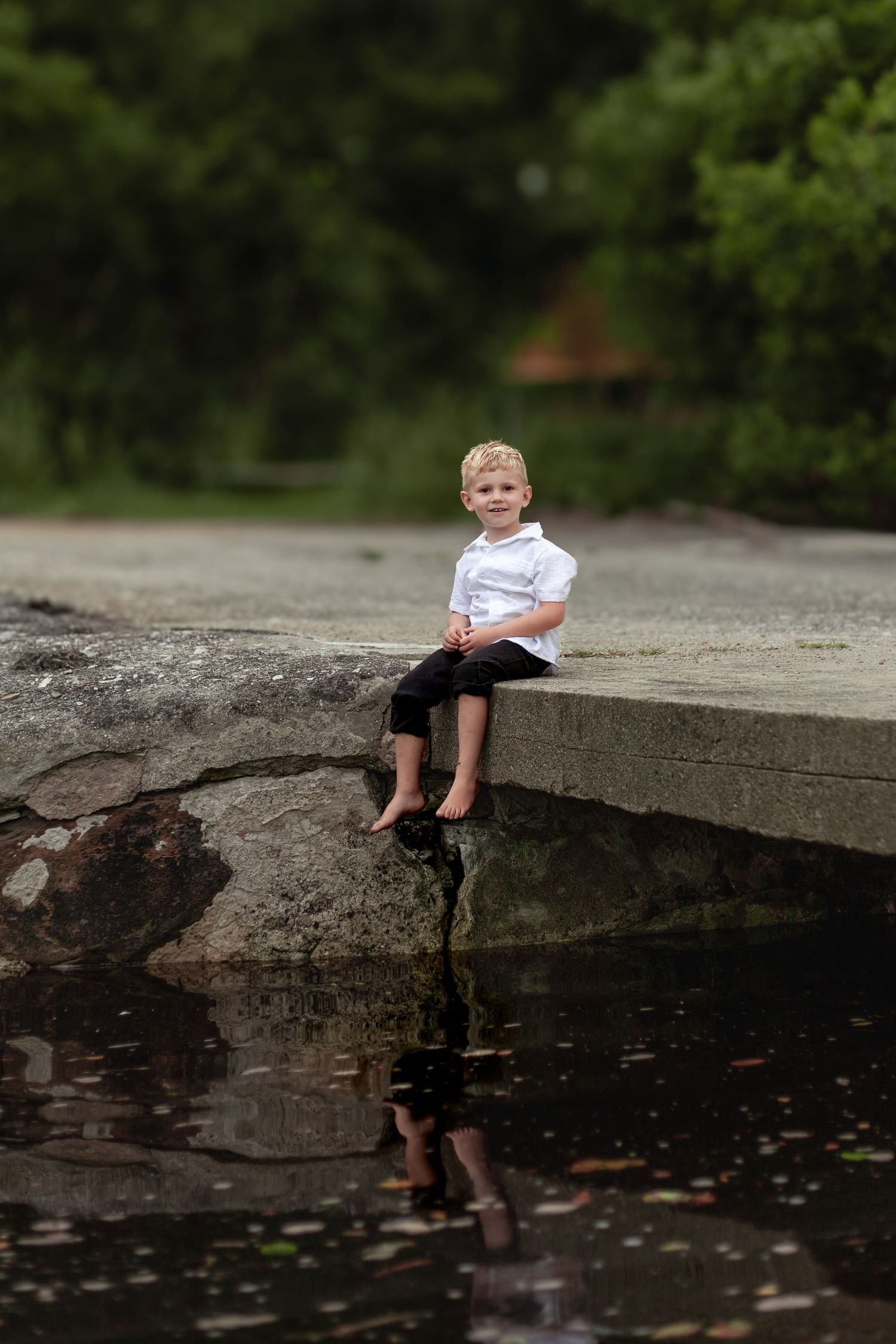 A child in a white shirt sits on a concrete ledge over a dark reflective pond surrounded by trees.