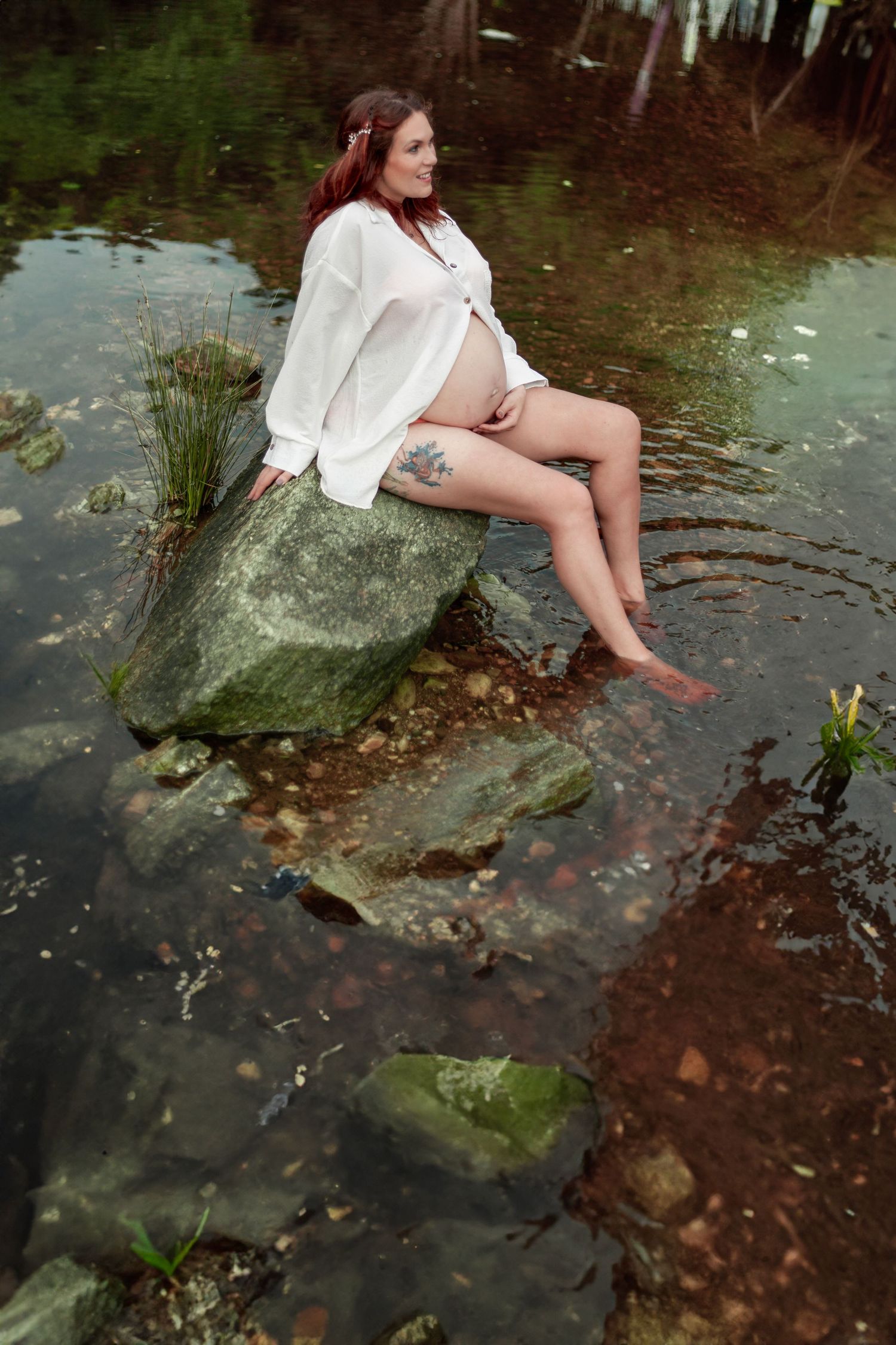 A person in a white shirt dress sits on a mossy rock by the water in a moody forest setting.