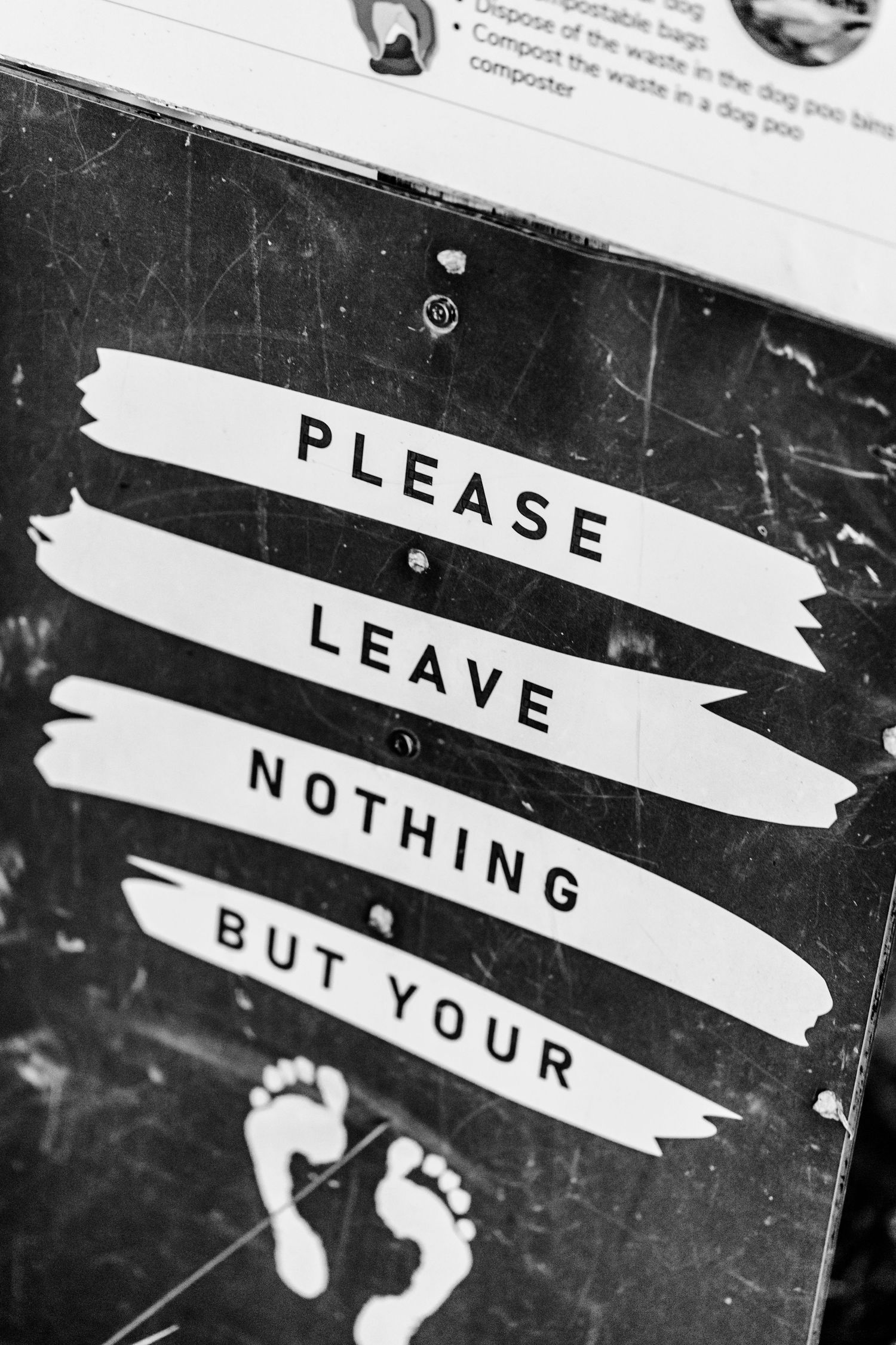 Black and white photo shows phrase 'PLEASE LEAVE NOTHING BUT YOUR' with footprint symbols on distressed surface.