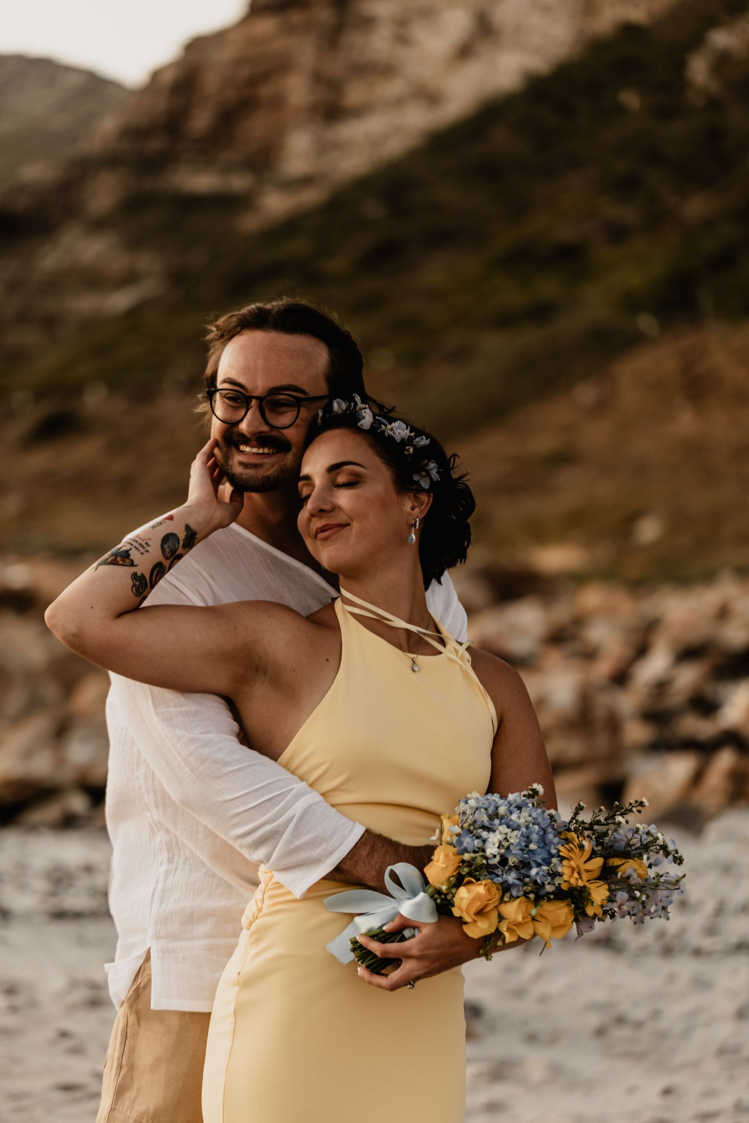 A romantic couple embraces on a sandy beach with mountains in the backdrop at sunset, carrying a blue and yellow bouquet.