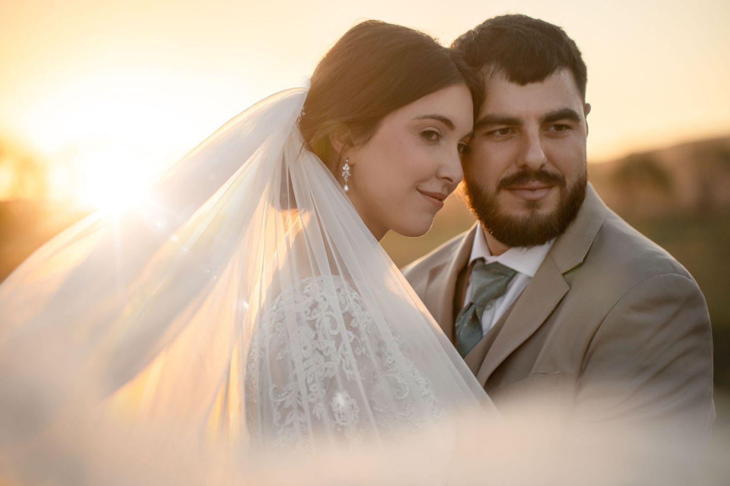 Bride and groom share a romantic golden hour sunset portrait, with flowing veil and warm glowing light.