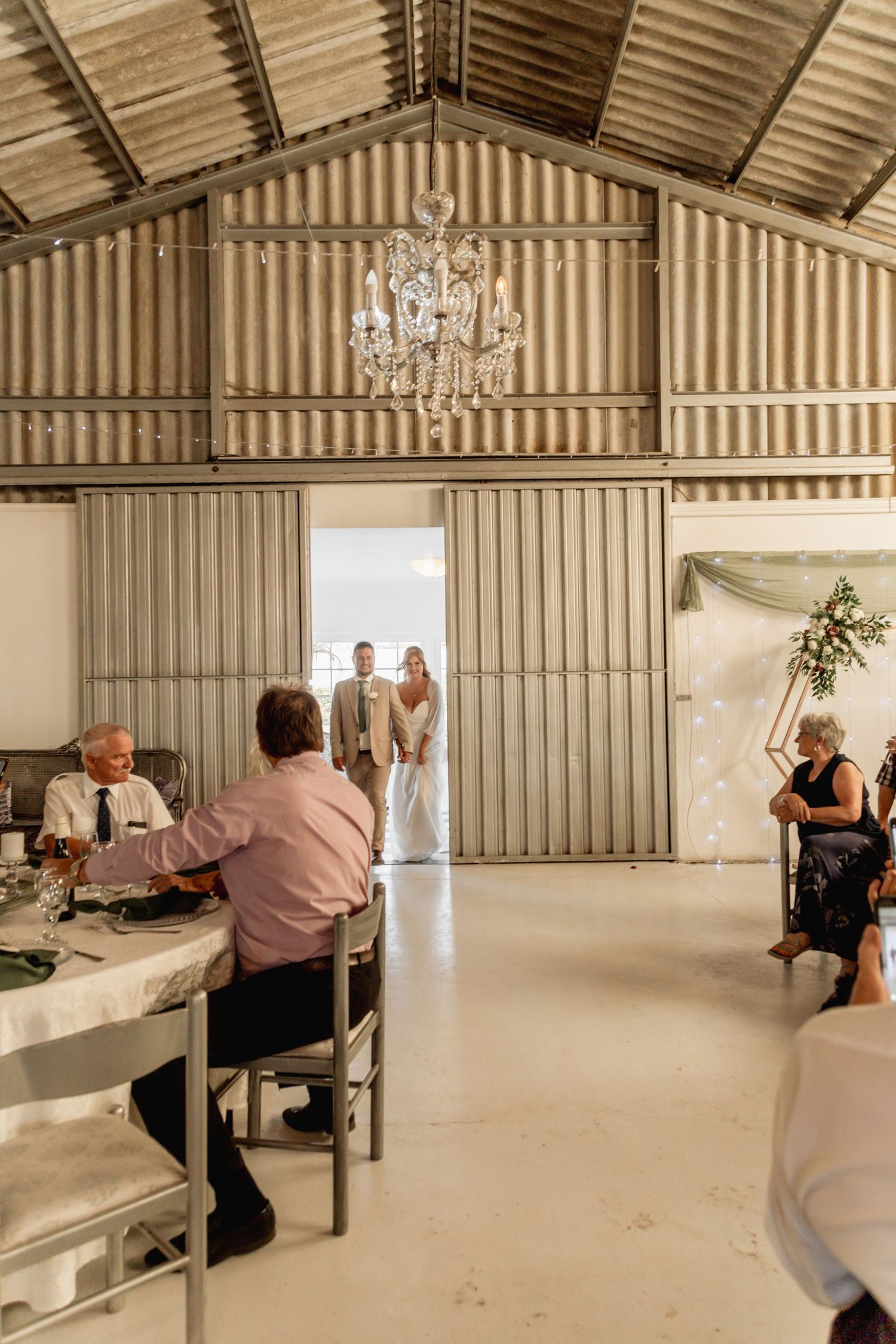 Rustic barn wedding reception with chandelier, corrugated metal walls, guests seated at tables as couple enters through large sliding doors.