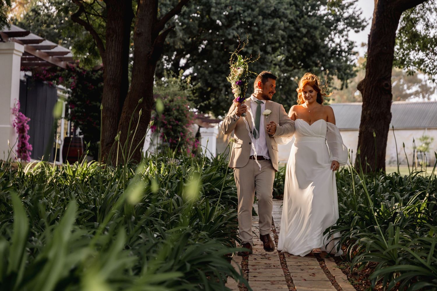 Bride and groom walking hand in hand through a lush garden pathway on their wedding day.