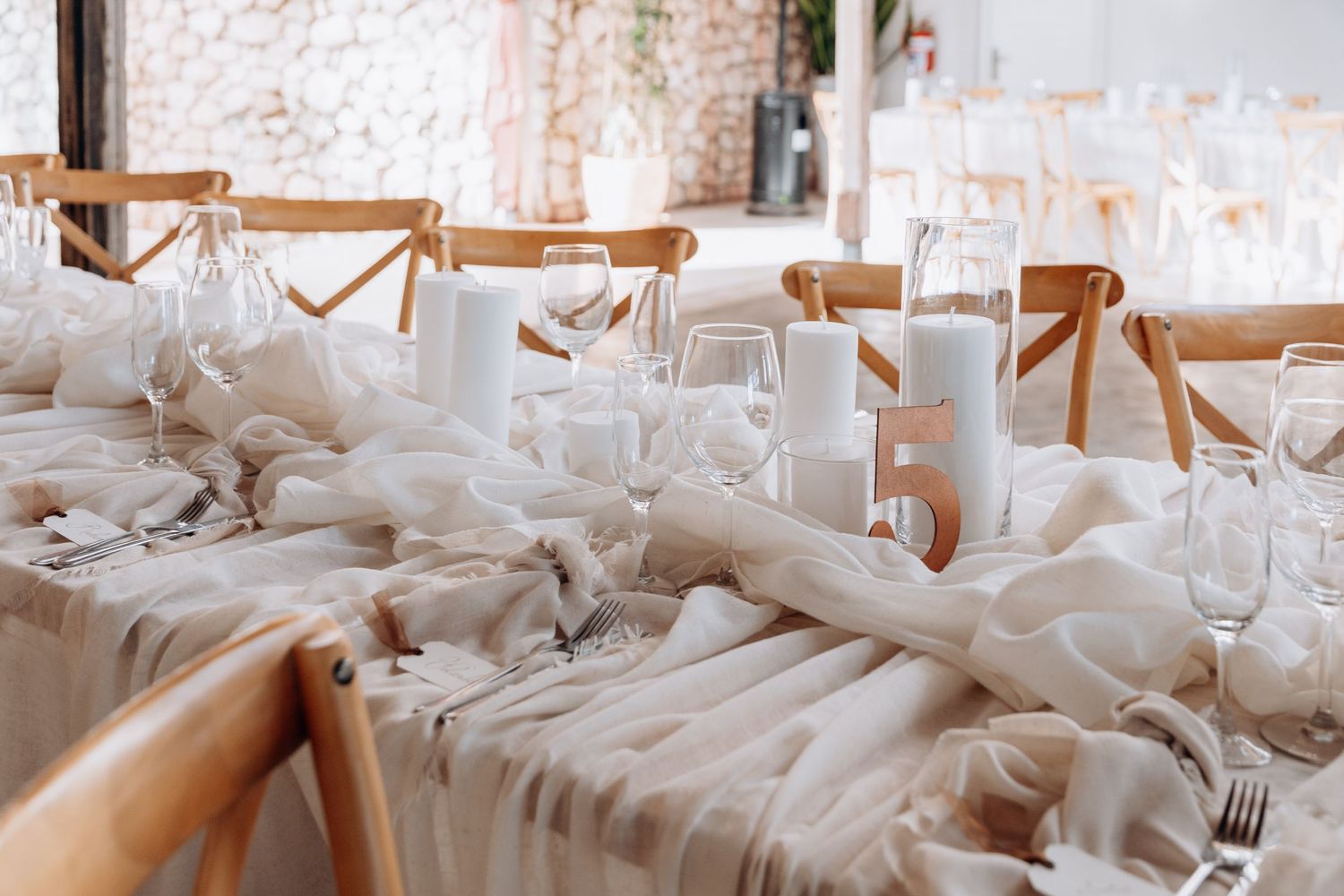 Elegant wedding reception table with white candles, draped fabric, and wooden chairs in a bright venue.