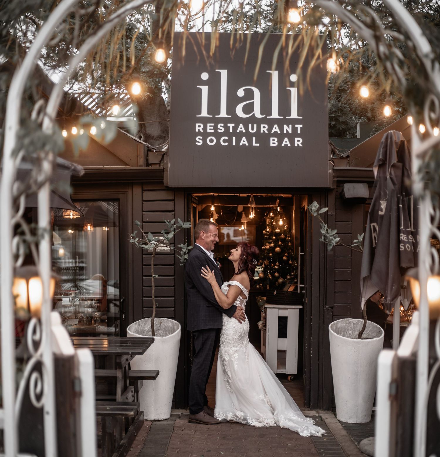 A romantic wedding couple shares a kiss at a dimly lit restaurant venue adorned with string lights and elegant decor.