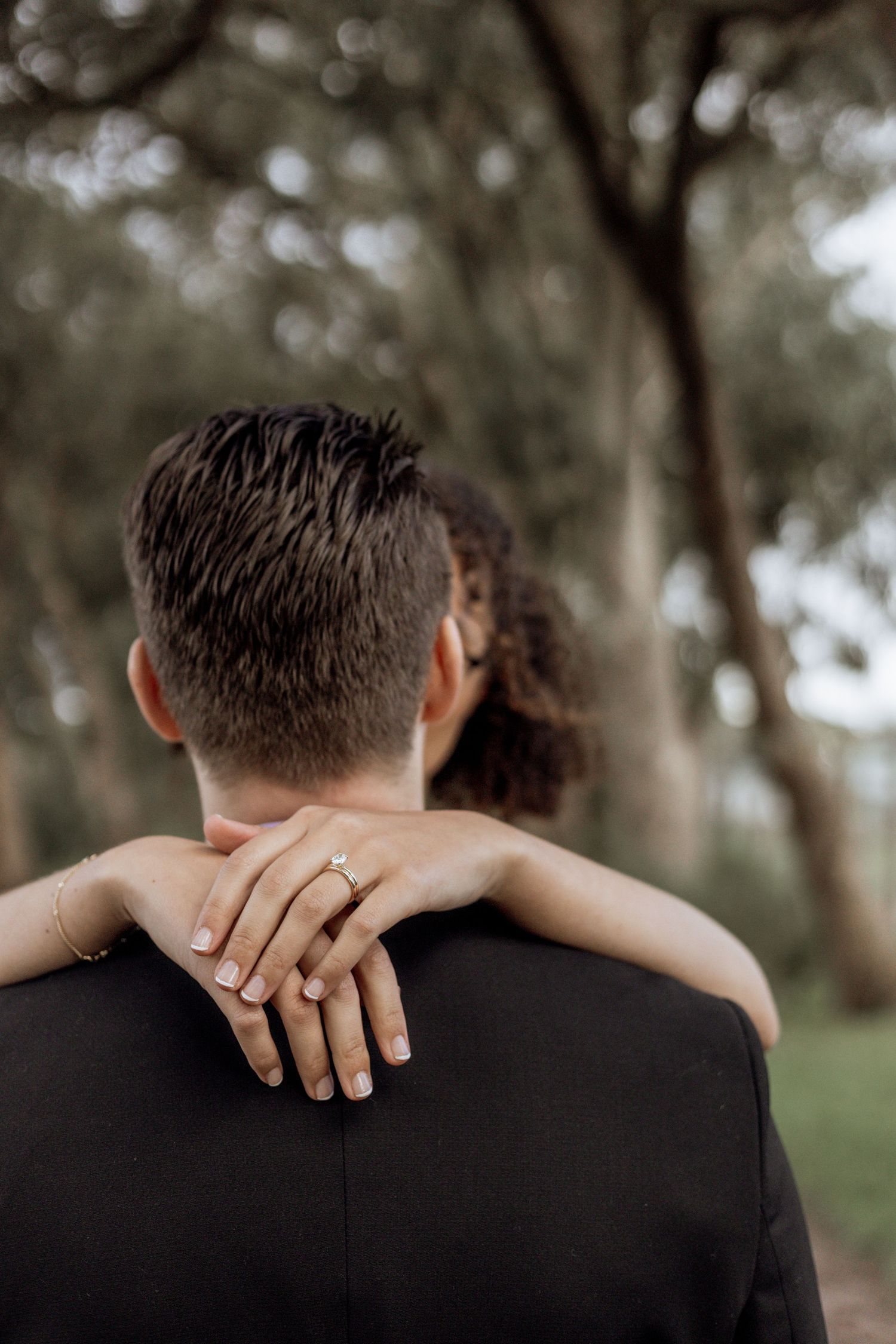 Close-up romantic shot shows interlocked hands embracing around neck against a blurred outdoor background.