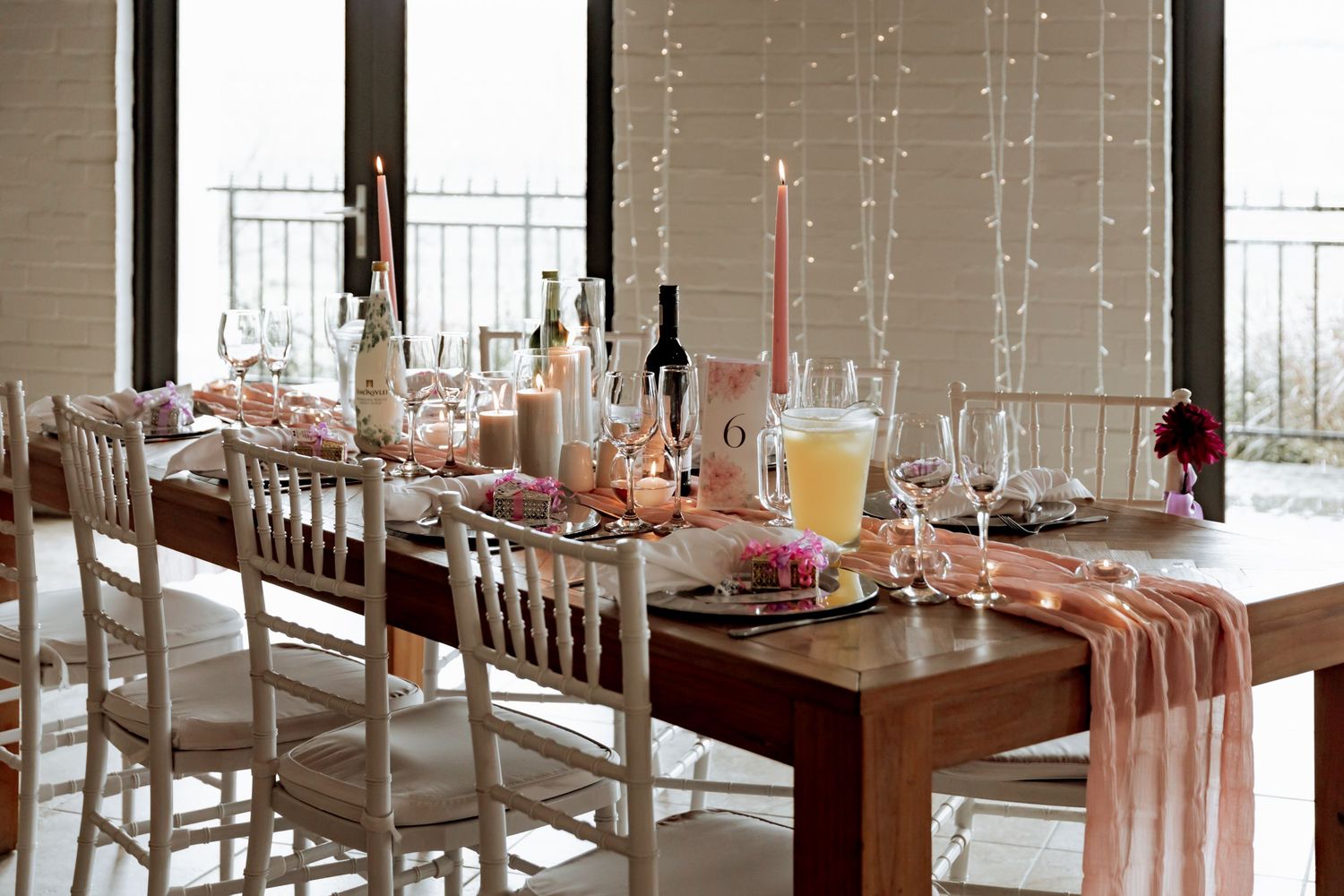 Elegant restaurant interior with long dining tables set with white linens, candles and wine bottles beside large windows.