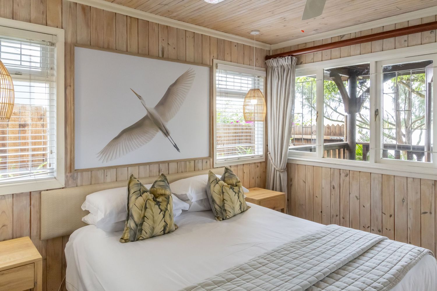 Cozy beach house bedroom with wood paneled walls, white bedding, and modern coastal decor featuring palm tree views.