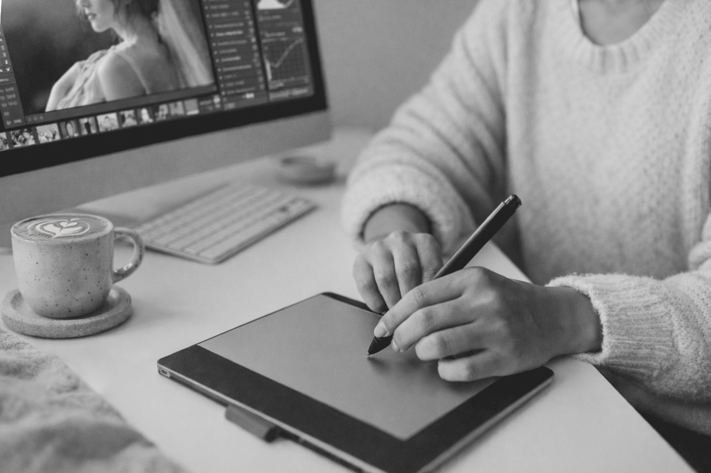 Someone works on a digital drawing tablet at a desk with a coffee cup and computer monitor in black and white.