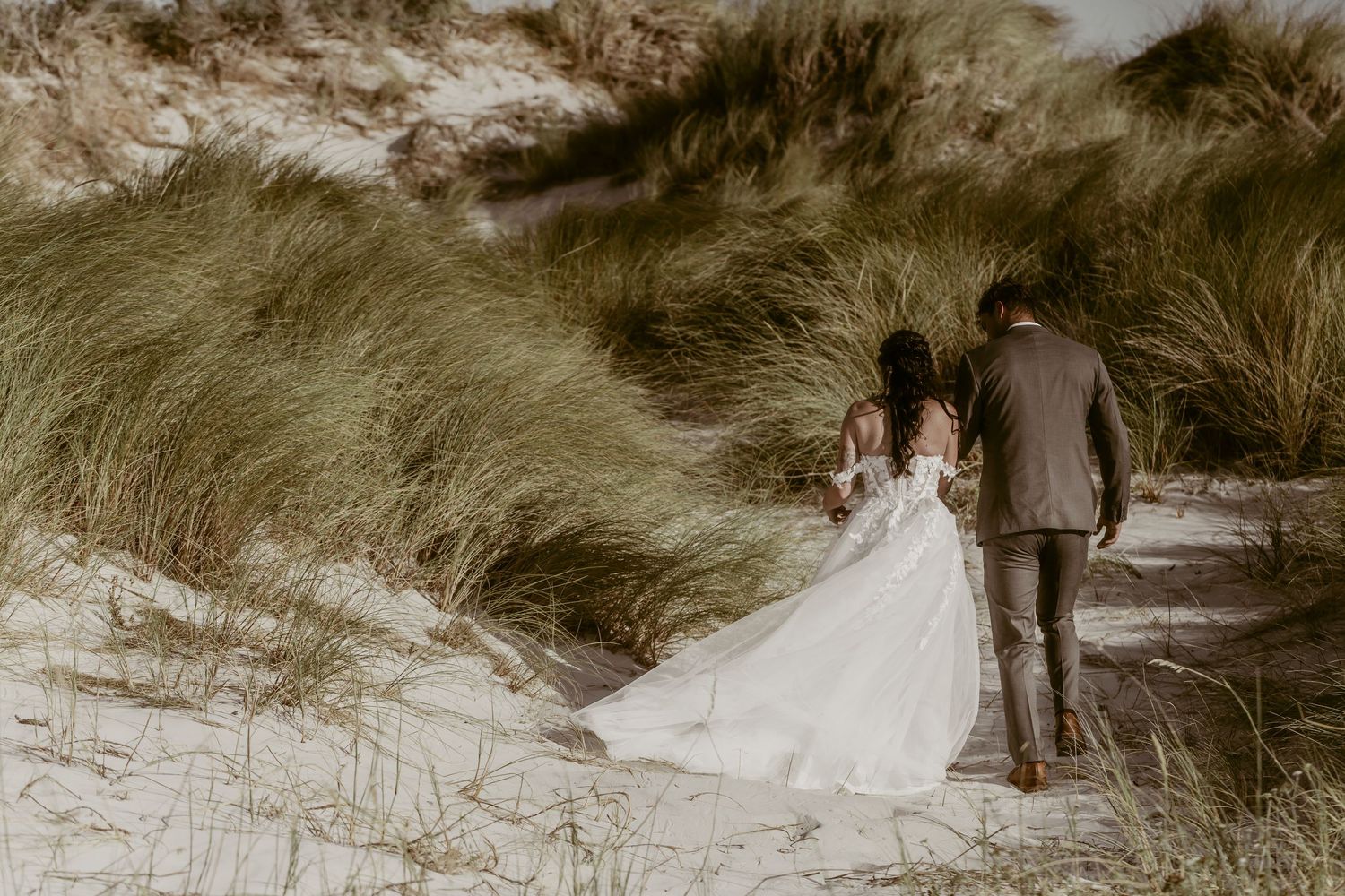 A bride in a flowing white gown and groom in a suit walk hand in hand through sandy dunes with tall grass.