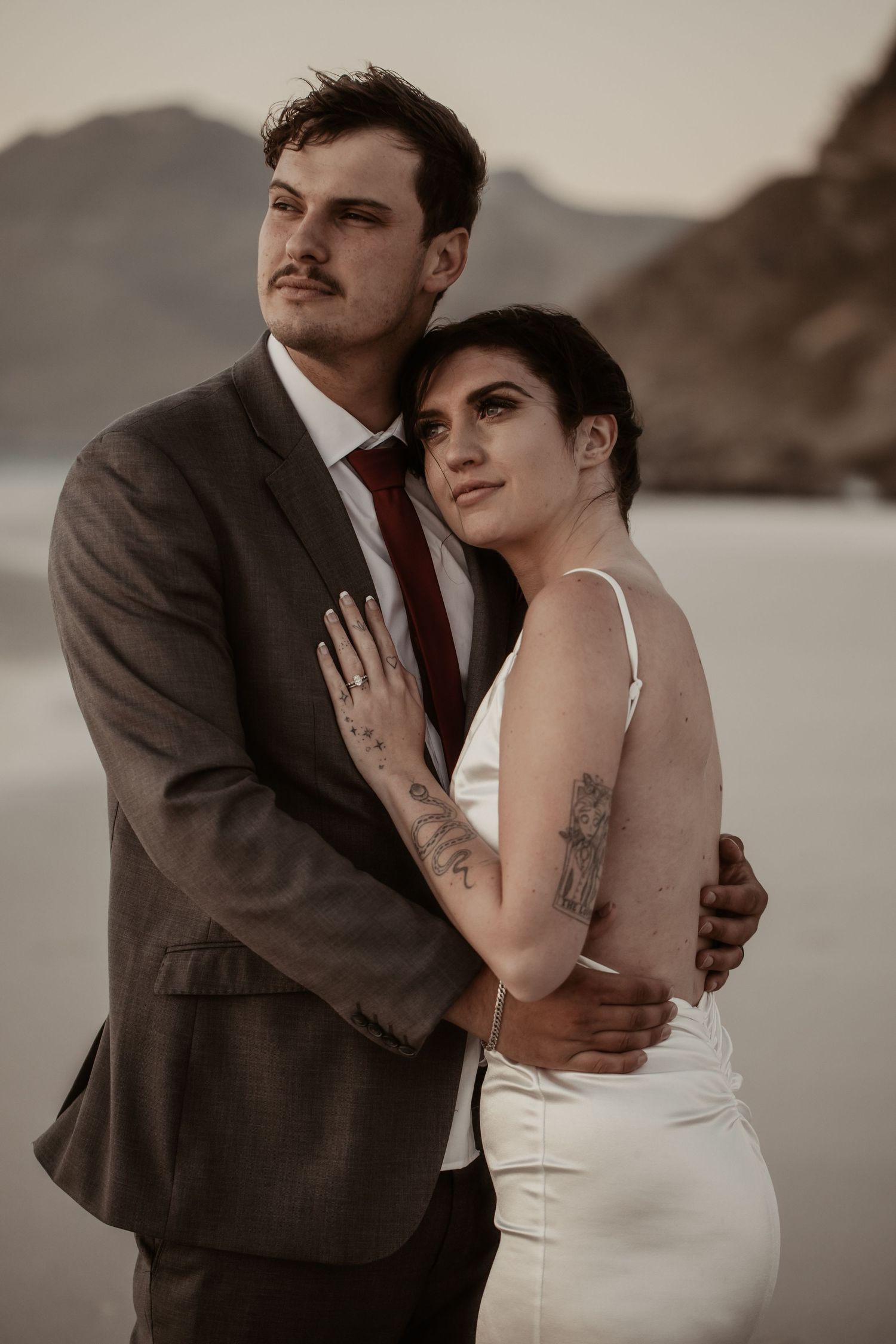 Couple embracing on beach, groom in gray suit with red tie, bride in white dress with tattoos.