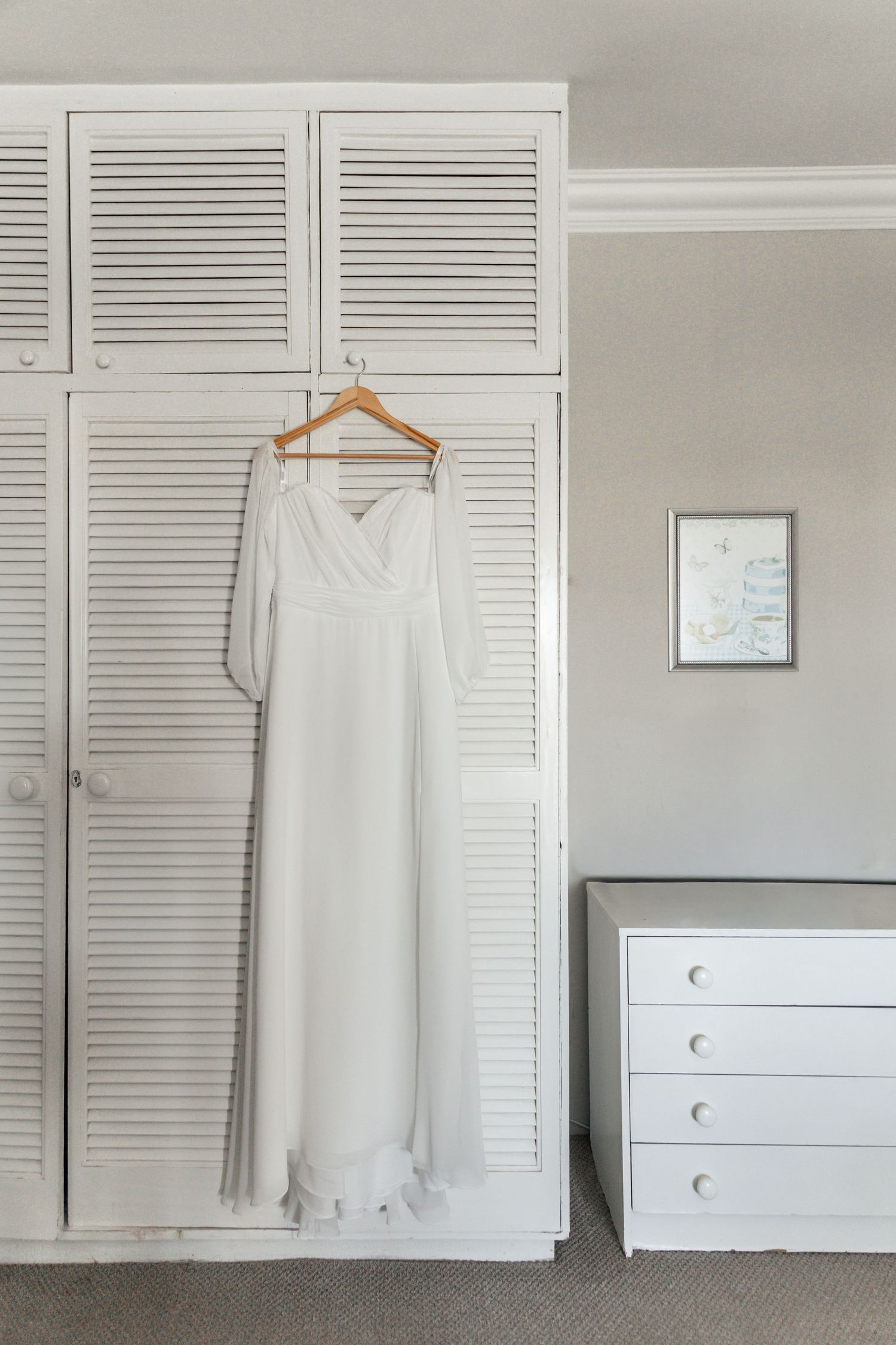 White wedding dress hanging on wooden hanger against white louvered wardrobe doors in bright bedroom.