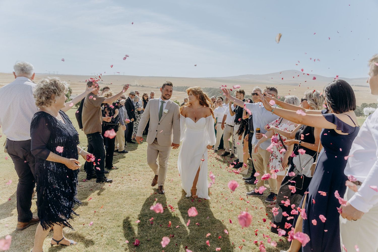 Newlyweds walk through rose petal confetti shower surrounded by cheering wedding guests outdoors.