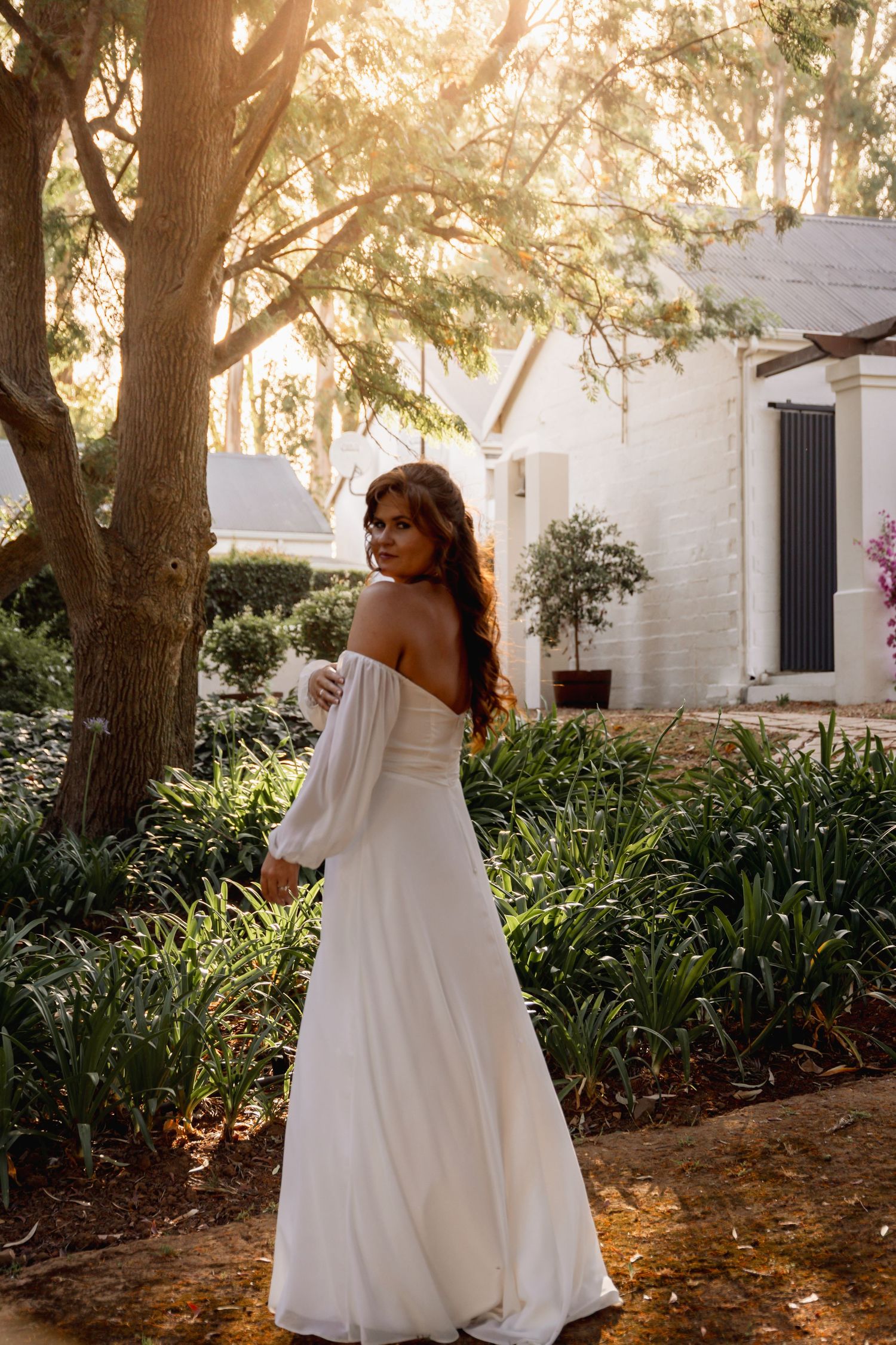 Bride in white off-shoulder gown with long sleeves poses in sunlit garden beside a tree near a white building.