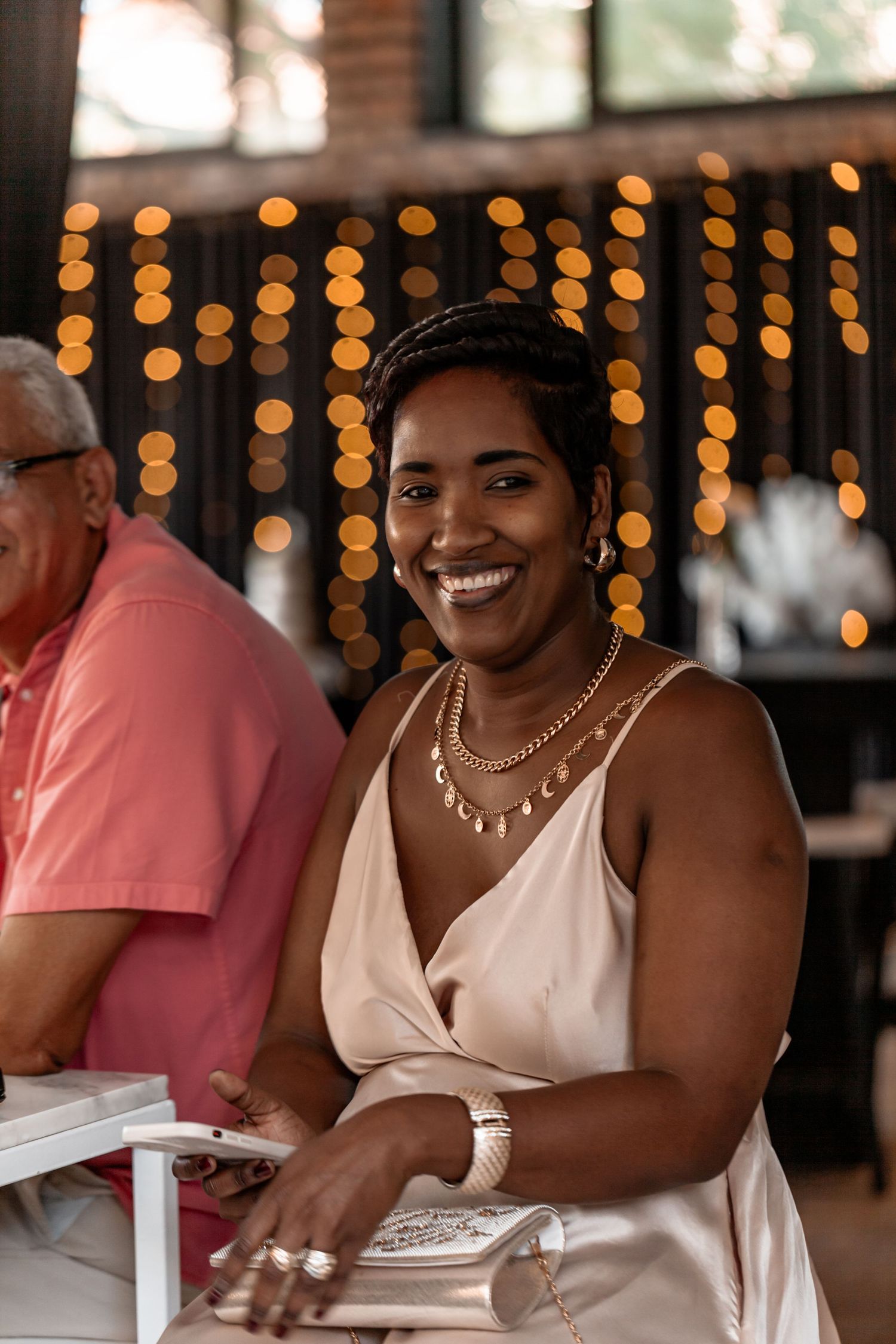 A person in a white dress smiles warmly as they sit at a table with soft bokeh lights in the background.