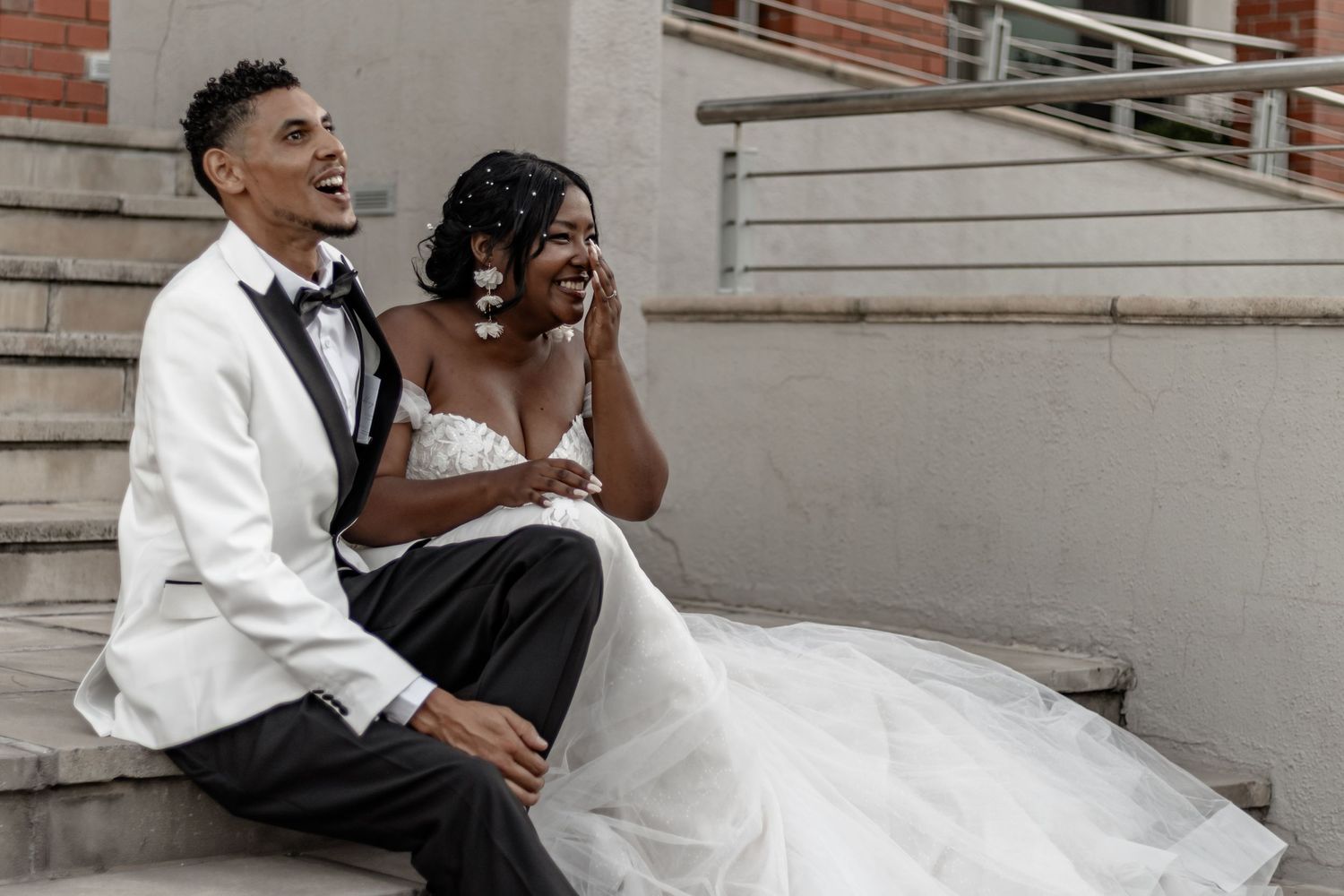 Wedding couple laughs joyfully while posing against grey wall at their celebration.