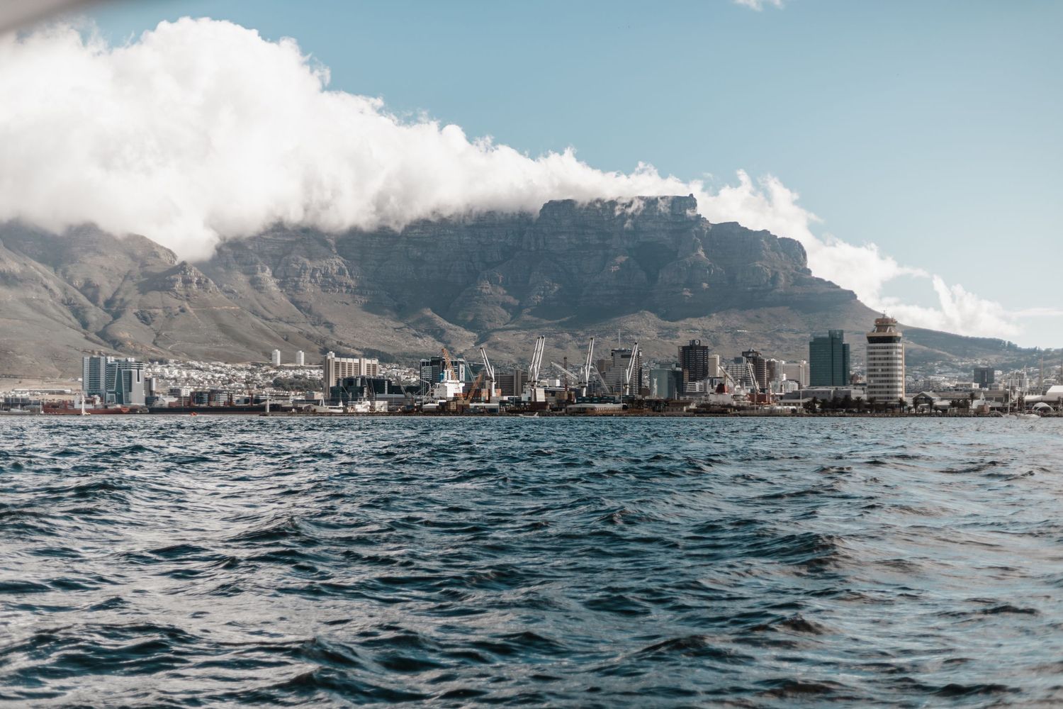 Coastal cityscape of Cape Town with dramatic Table Mountain backdrop and blue ocean waters in the foreground.