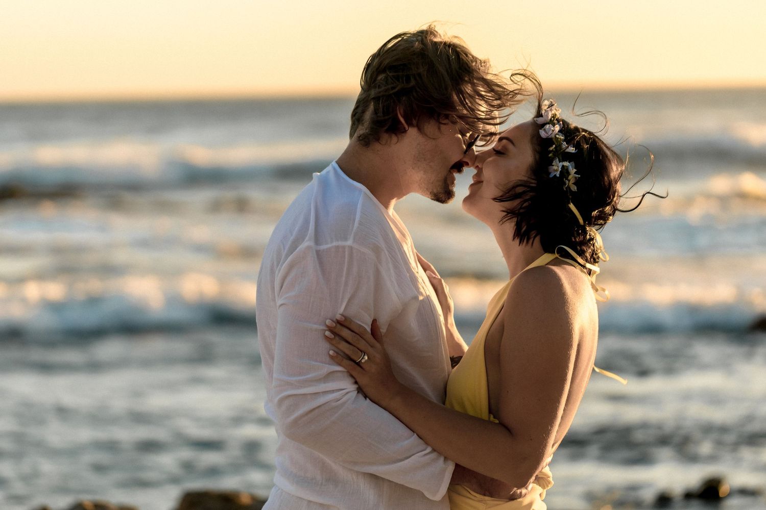 A romantic couple shares a tender kiss on the beach at sunset with ocean waves in the background.