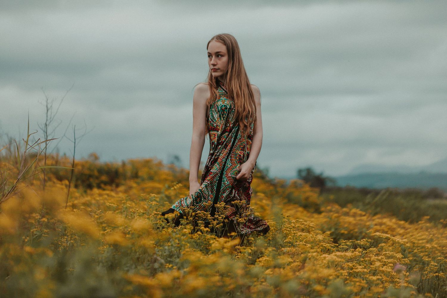 A figure sits among vibrant yellow wildflowers in a moody field beneath overcast skies.