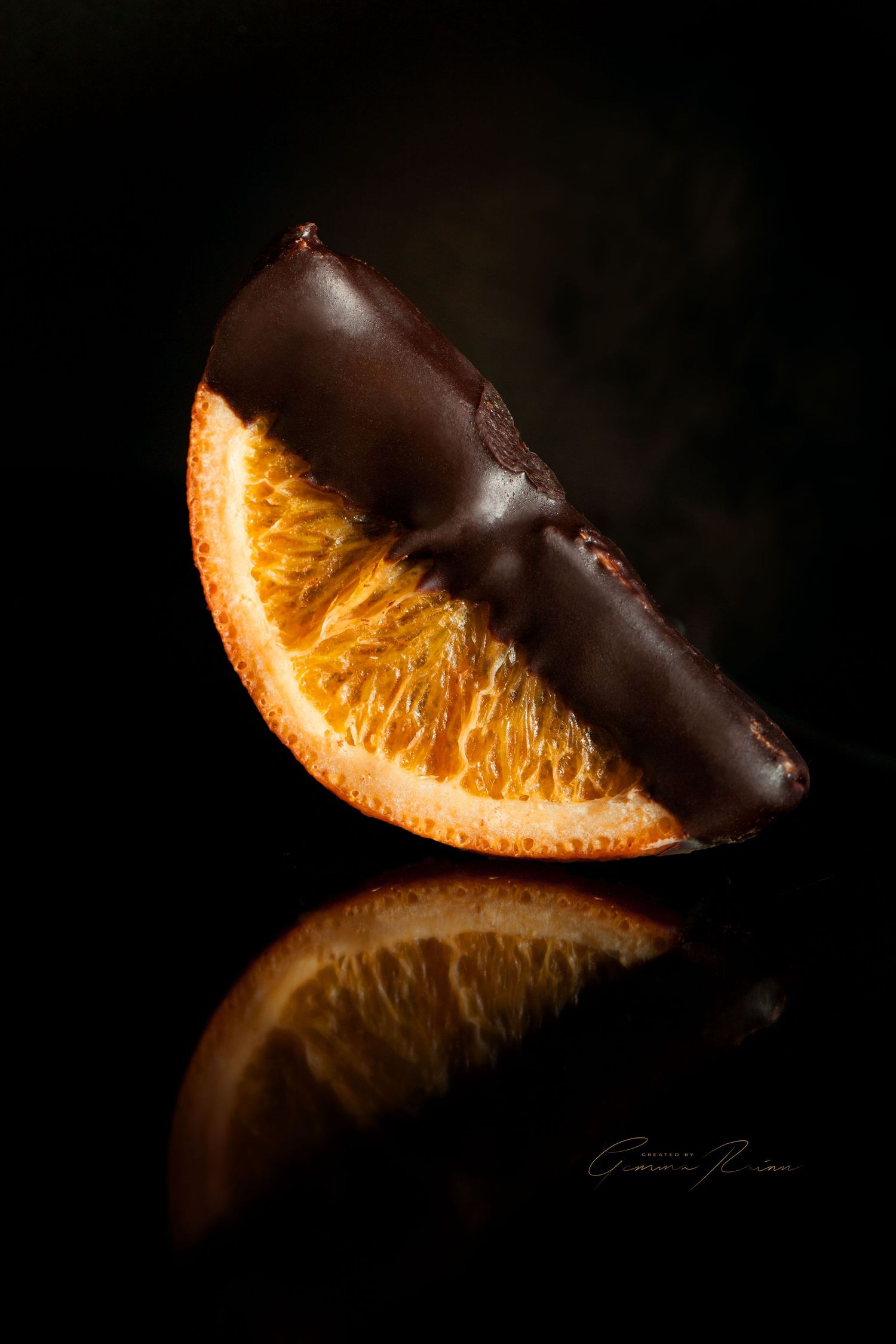 Dark chocolate dipped orange slices arranged artistically on black reflective surface with dramatic lighting.