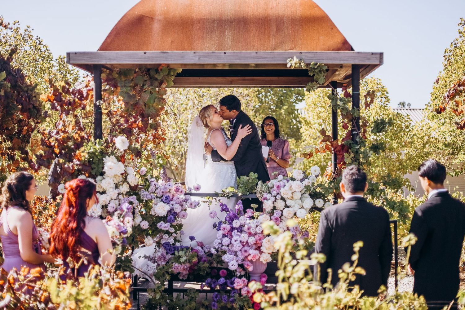 Couple shares first kiss under floral gazebo at outdoor wedding ceremony surrounded by purple and white blooms.