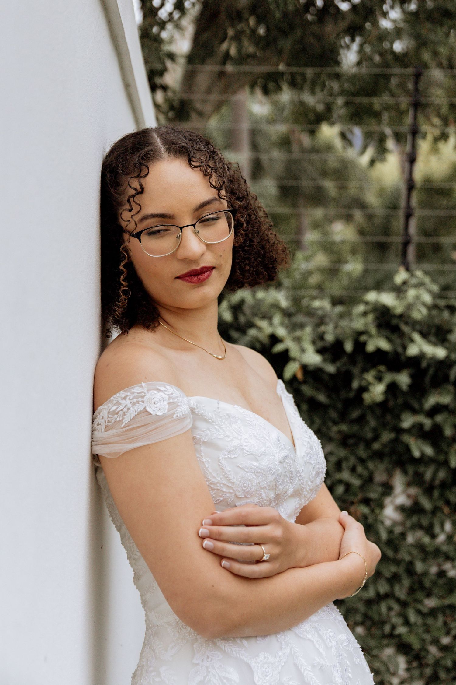 Close-up portrait of a bride in a delicate lace off-shoulder wedding gown with glasses against a green garden backdrop.