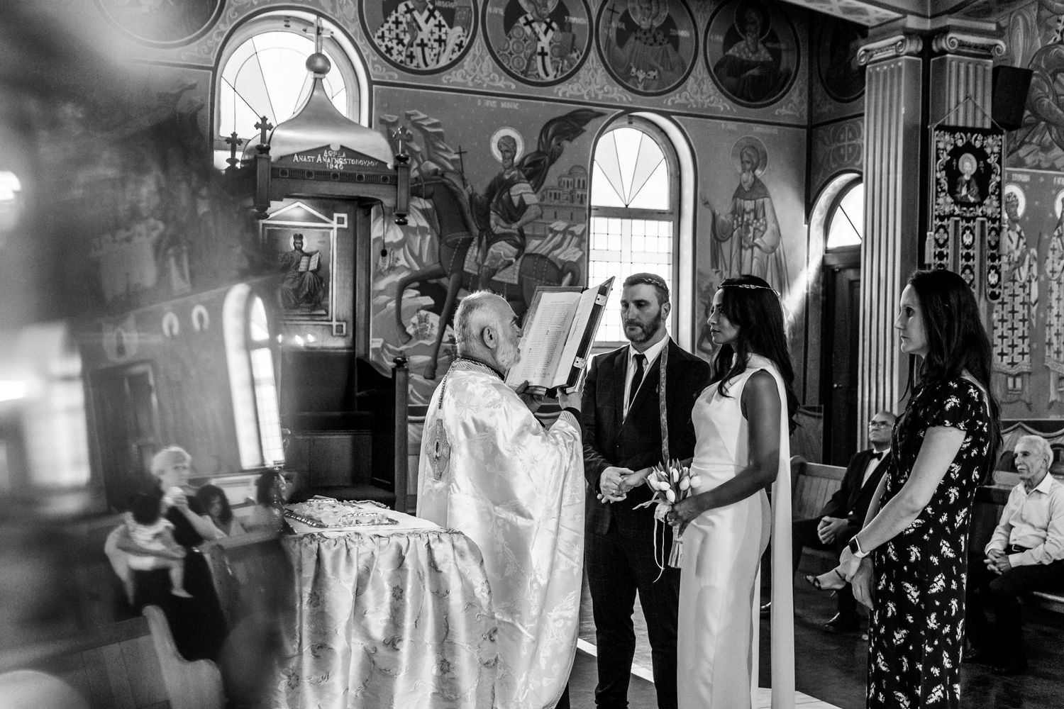 Black and white photo of a couple exchanging vows in an Orthodox church ceremony with a priest officiating.
