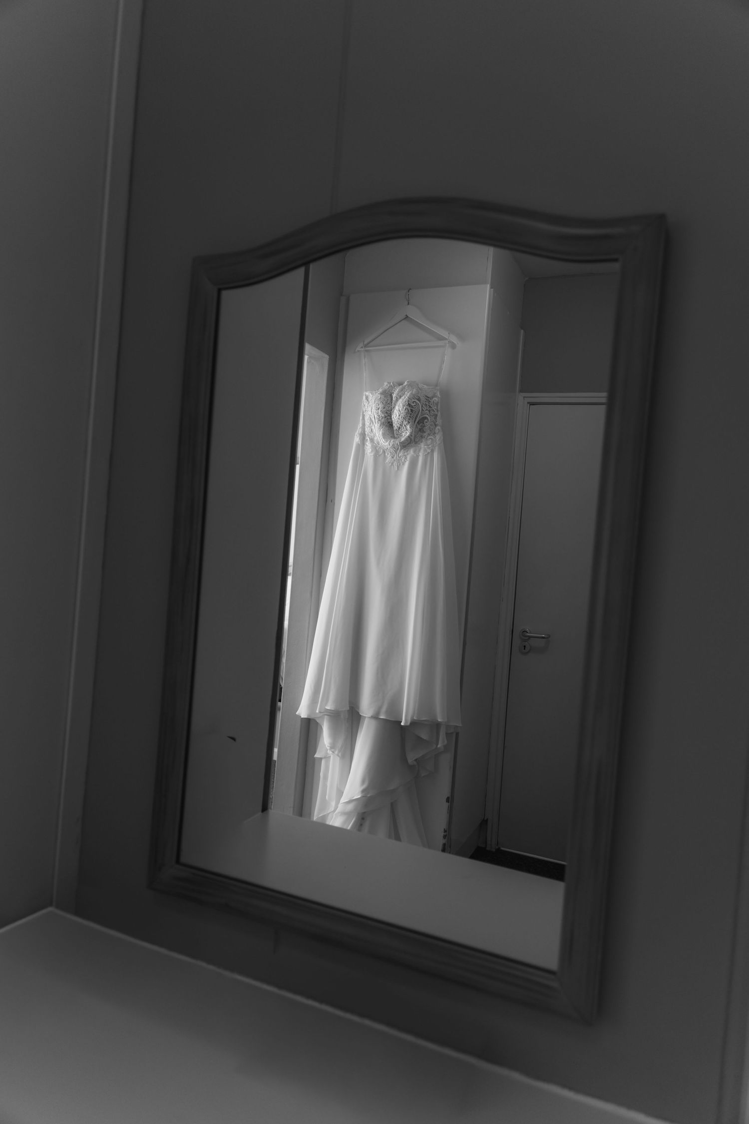 Black and white photo of a wedding dress hanging on a hanger, reflected in an ornate framed mirror.